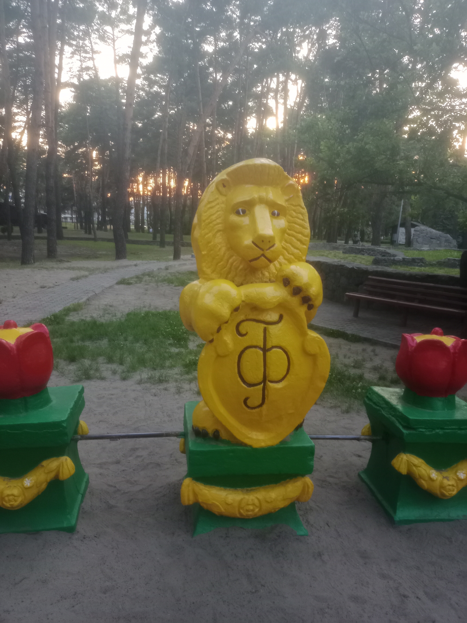 The image shows a statue of a yellow lion holding a shield with a Cyrillic letter "Ф" inscribed on it. The lion is standing on a green base, which is decorated with yellow floral motifs. Flanking the lion are two green pedestals, each topped with a red tulip sculpture. A metal bar connects the two pedestals. The scene is set outdoors in a park-like environment, with tall trees in the background and a sandy ground. The sun is setting, casting a warm glow through the trees. In the background, there are paved paths, a wooden bench, and some stone structures. The overall atmosphere is serene and somewhat whimsical. This statue is located in the city of Horishni Plavni, Ukraine, likely in a public park or recreation area.