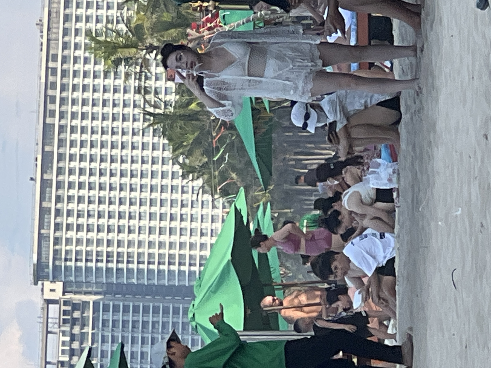 The image depicts a crowded beach scene in Da Nang, Vietnam, with a large, modern hotel building visible in the background. The hotel is a multi-story structure with numerous windows and balconies, suggesting it's a prominent landmark. In the foreground, people are gathered on the sand, with several green umbrellas providing shade.
Several individuals are the focus of the image. A woman in a white lace bikini and cover-up is prominently positioned, holding a phone to take a picture of herself. She is wearing glasses and her hair is styled in a bun. To her left, a person wearing a white hat and mask is seated, looking at their phone. To the left of the woman in white, a man in a green shirt and dark pants is speaking to someone, gesturing with his hand. Further into the scene, several other individuals are seated or standing, some interacting with each other, while others are engaged with their phones. Beach umbrellas, bags, and what appear to be pieces of colorful fabric strung between poles are also visible, indicating a festive or organized beach setting. The overall atmosphere appears casual and summery, with a mix of locals and tourists enjoying the beach.
