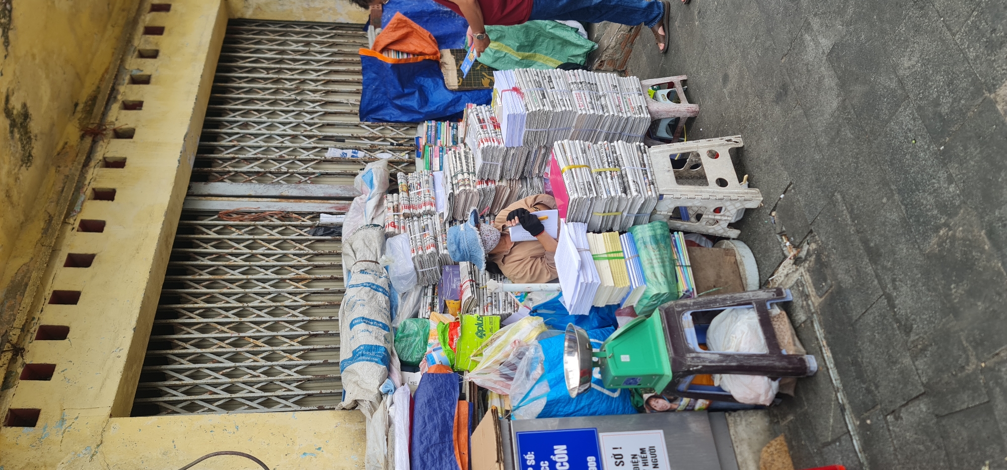 This outdoor image captures a vibrant street scene, likely a vendor's stall, in Đà Nẵng, Vietnam. The main subjects are two individuals and an extensive collection of stacked papers and newspapers.
One person, seated amidst the bundles, wears a light brown long-sleeved top, dark gloves, and a hat, seemingly engrossed in writing or taking notes on a piece of paper with a pen. They are surrounded by towering stacks of neatly bundled newspapers, secured with yellow and white ties, along with piles of various colored blank papers (white, yellow, green, blue). Another person is partially visible standing at the top right, wearing a red shirt, blue jeans, and sandals, appearing to interact with the stall or perhaps passing by.
The scene is set alongside a faded yellow building featuring a closed, metallic rolling shutter, common for storefronts. Along the top of the wall, a row of small, square, reddish-brown indentations is visible. The ground is a worn, gray paved sidewalk. Various objects are scattered around the stall, including several plastic stools of different colors (white, gray, brown), blue and orange tarps, numerous plastic bags, and a green and silver weighing scale. A box with "Aplus" printed on it is also seen.
The activity suggests a bustling commercial spot, possibly a newsstand, a lottery ticket vendor, or a collection point for recycled paper. The person taking notes, surrounded by bundles of paper and a scale, implies transactions or inventory management.
Visible text on a blue sign or box at the bottom right includes "SỐ!" (likely "Number!" or "Issue!"), partial words like "ĐIỆN", "NGƯỜI", and "CON", and "009", suggesting it could be related to lottery tickets or other numbered items. The overall impression is one of everyday commerce in an urban Vietnamese setting during daylight hours.