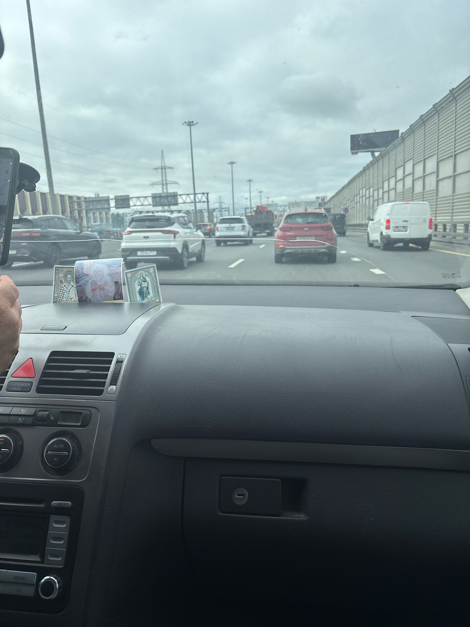 A first-person perspective shot from inside a car shows a heavily trafficked highway with multiple lanes of vehicles. The sky is overcast. In the foreground, on the dashboard, there are religious icons and currency. The car's interior, including the steering wheel and center console, is visible. The road ahead is lined with sound barriers, and power lines and utility poles are visible in the distance.