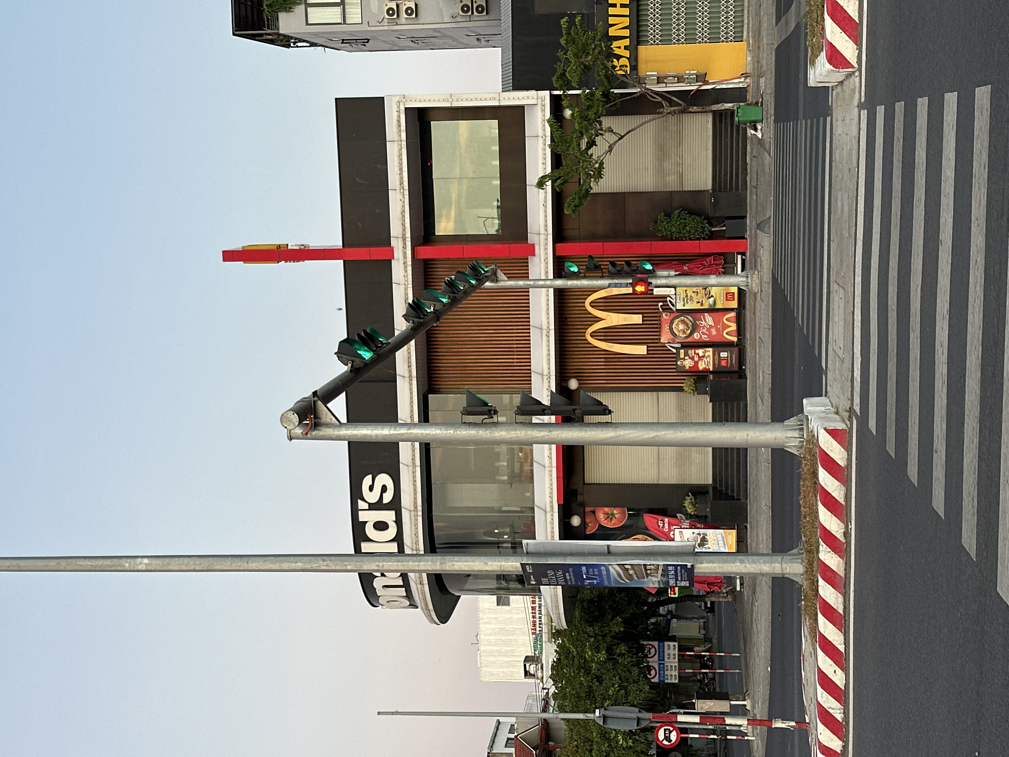A McDonald's restaurant is visible on the corner of an intersection in Da Nang, Vietnam. The restaurant has a large, illuminated golden arches logo and a black facade. In front of the restaurant, a pedestrian crossing is marked on the asphalt street, bordered by red and white striped concrete barriers. Traffic lights and streetlights are also present. The sky is a clear, pale blue, suggesting it is either early morning or late afternoon. Several trees are planted along the sidewalk. The street appears to be a commercial area, with other buildings and storefronts visible in the background.