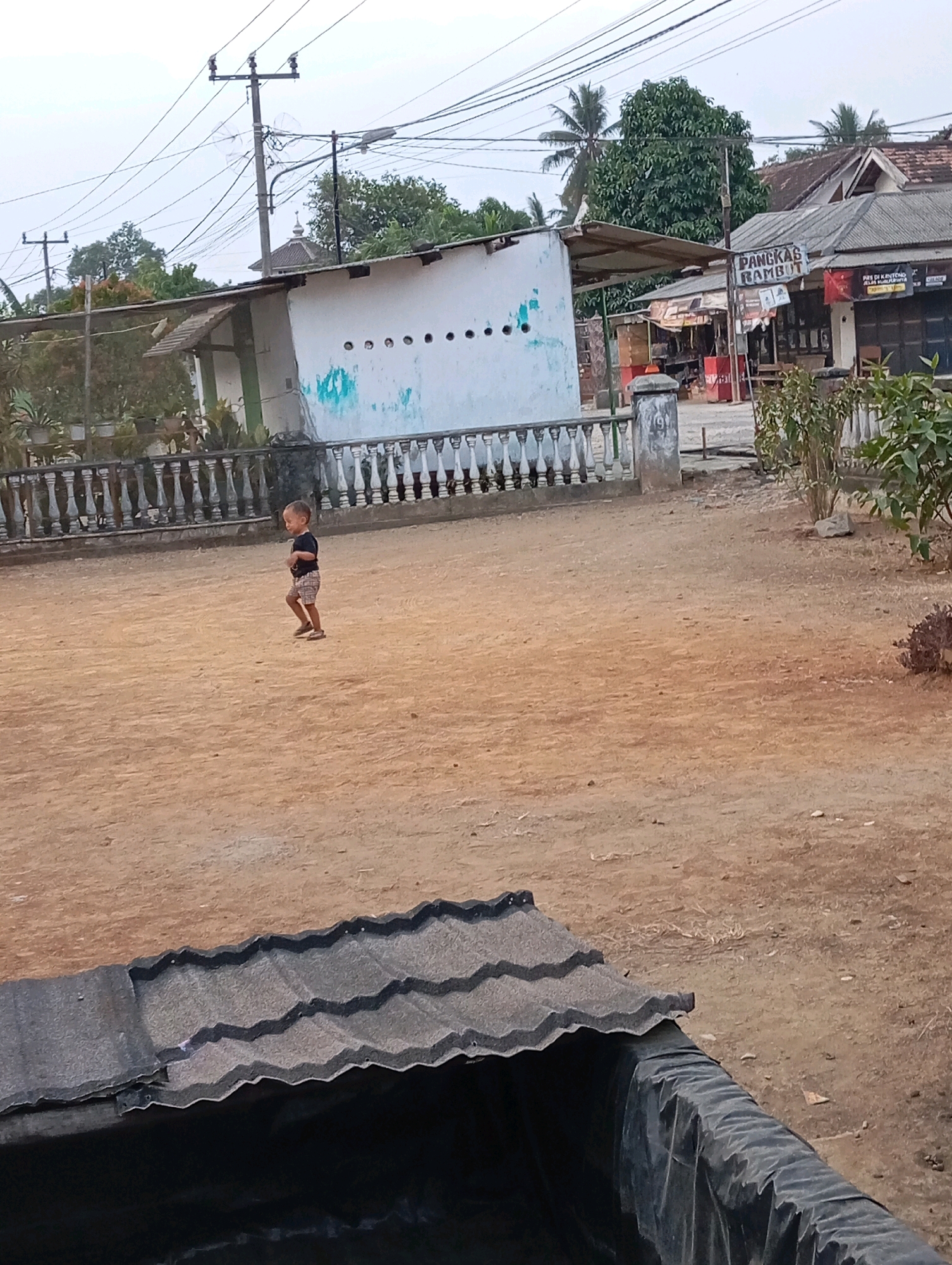 The image depicts a scene in Leman Benawi, Indonesia, featuring a young boy in the foreground, standing in a dirt-covered yard. The boy, wearing a dark t-shirt and plaid shorts, is looking towards the left of the frame and appears to be in motion. Behind him, a white building with a row of circular openings is visible, bordered by a decorative balustrade. To the right of the boy, there is a building with a sign that reads "PANGKAS RAMBUT," indicating a barbershop. Further in the background, electrical poles and wires crisscross the sky, and lush green trees dot the landscape. In the bottom foreground, a portion of a black tarp and a gray, corrugated roof are visible, suggesting some sort of construction or outdoor covering. The overall environment appears to be a mix of residential and commercial spaces, typical of a small Indonesian town.