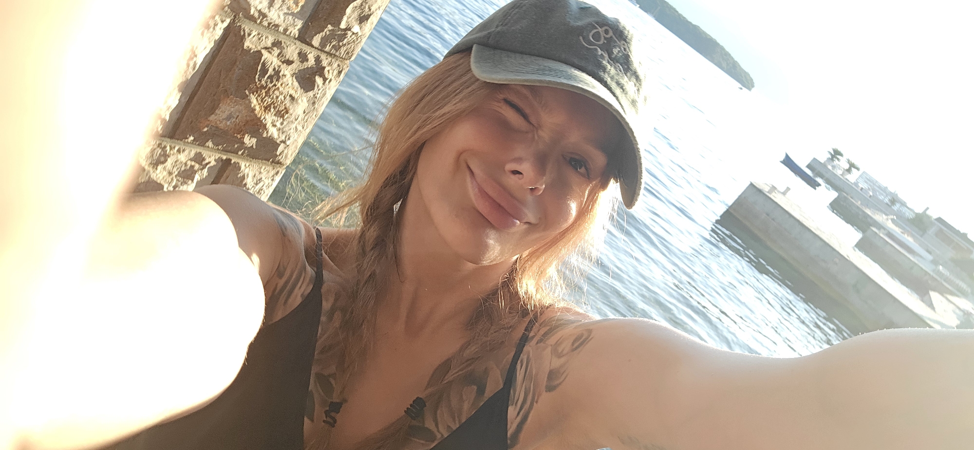 A selfie taken outdoors in bright sunlight. A fair-skinned woman with braided red-blonde hair and tattoos on her chest and arms winks at the camera. She wears a dark tank top and a weathered gray baseball cap with a logo. Behind her is a body of water, possibly the Adriatic Sea, and in the distance, a coastline. There is a stone wall to her left and a pier or dock-like structure to the right. The location is likely Bogišići, Montenegro.