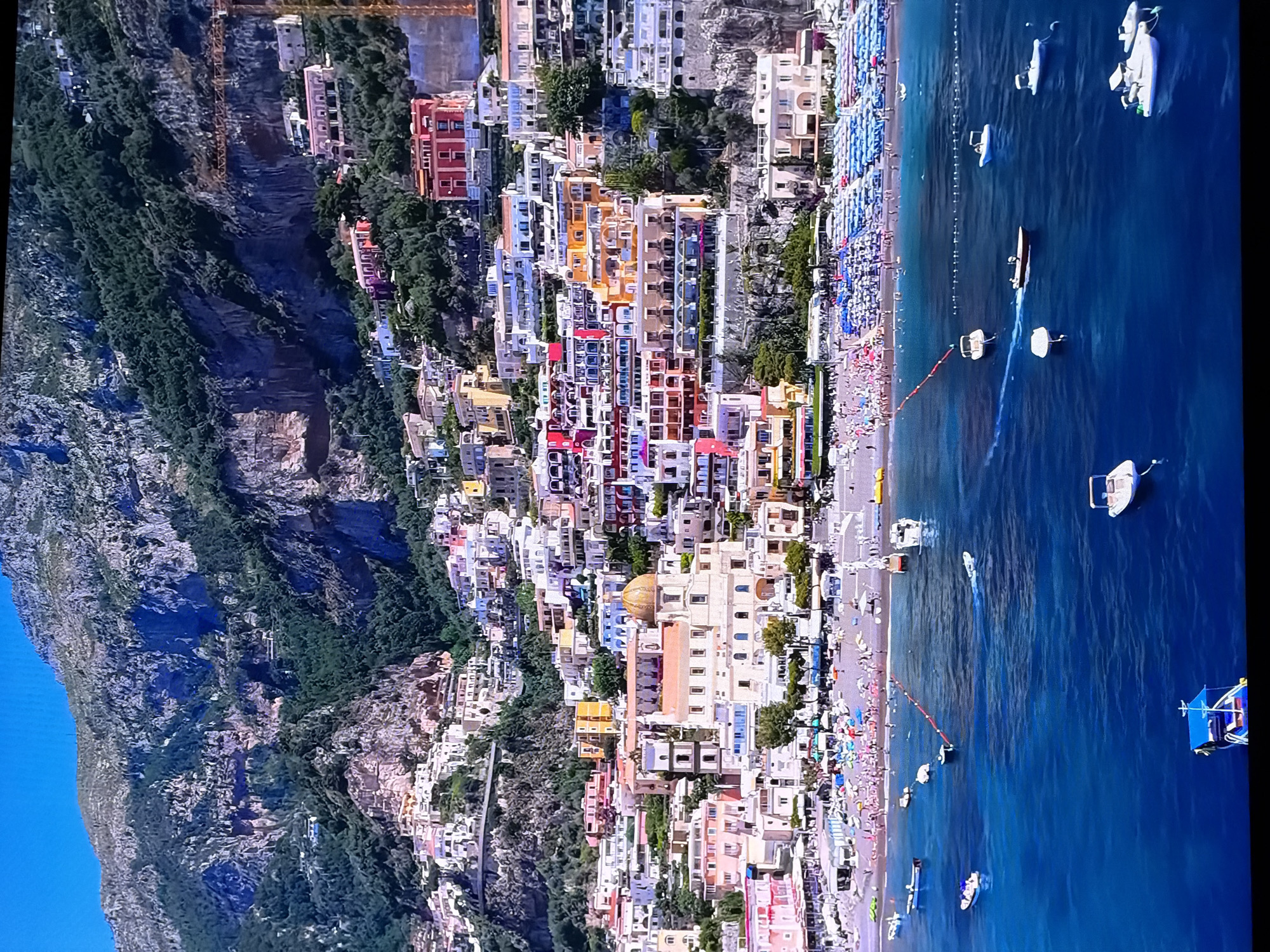 The image displays a vibrant, bustling scene in Positano, Italy, a picturesque town nestled on the Amalfi Coast. The characteristic colorful houses cascade down a steep cliff face, meeting a lively beach crowded with sunbathers and colorful umbrellas. The azure waters of the Tyrrhenian Sea are dotted with various boats, from small dinghies to larger motor yachts, some moored and others in motion. The background is dominated by the dramatic, rugged cliffs and lush greenery that define the Amalfi Coast landscape.
This image is not related to Chirchik, Uzbekistan.