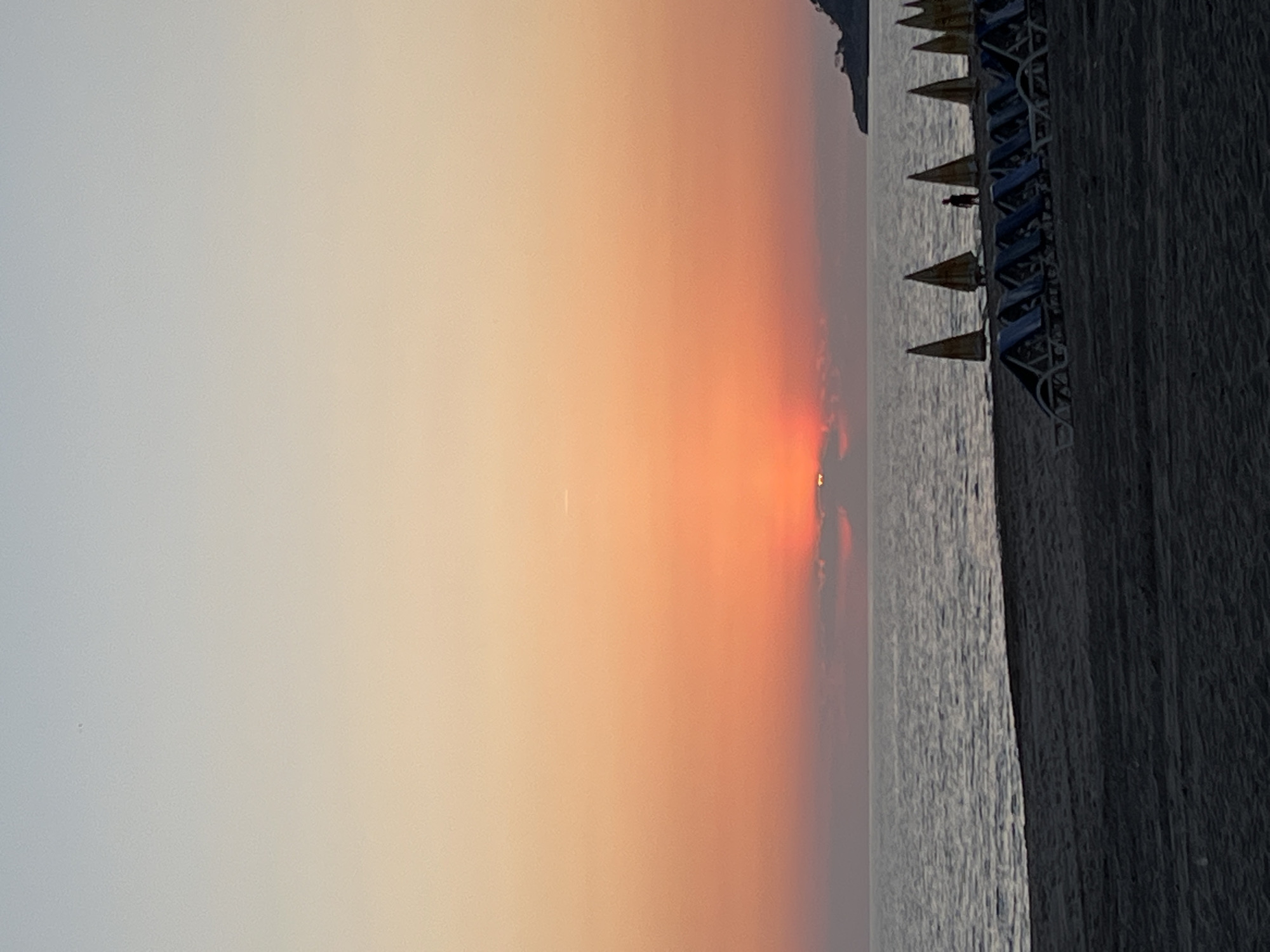 The image shows a sunset scene on a beach in Alanya, Turkey. The sky is a gradient of light gray to orange, with the sun partially obscured by clouds. A row of beach umbrellas and chairs lines the edge of the beach, with the sea in between. The beach itself is dark and appears to be composed of sand or pebbles. A small figure can be seen between the rows of umbrellas.