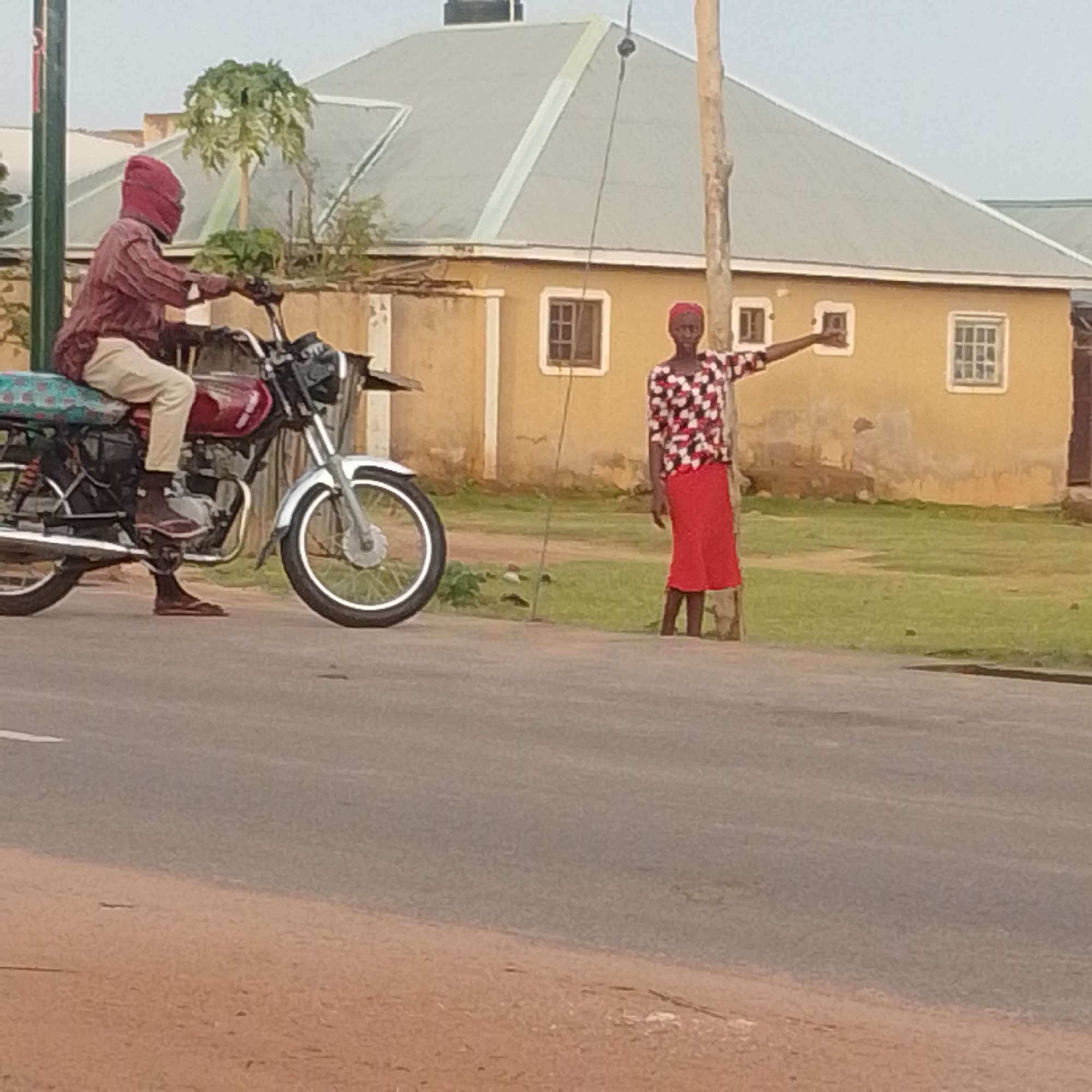 A man wearing a maroon shirt and beige pants, with a red head covering, rides a red motorcycle on a paved road. He is looking forward. To his right, a woman wearing a red skirt and a patterned top with red, white, and black colors is standing near a utility pole. Her right arm is extended outwards. Behind them, there is a tan-colored building with a grey roof and several windows. Green grass and some trees are visible in the background. The sky is overcast. The scene appears to be in Tempure, Nigeria.