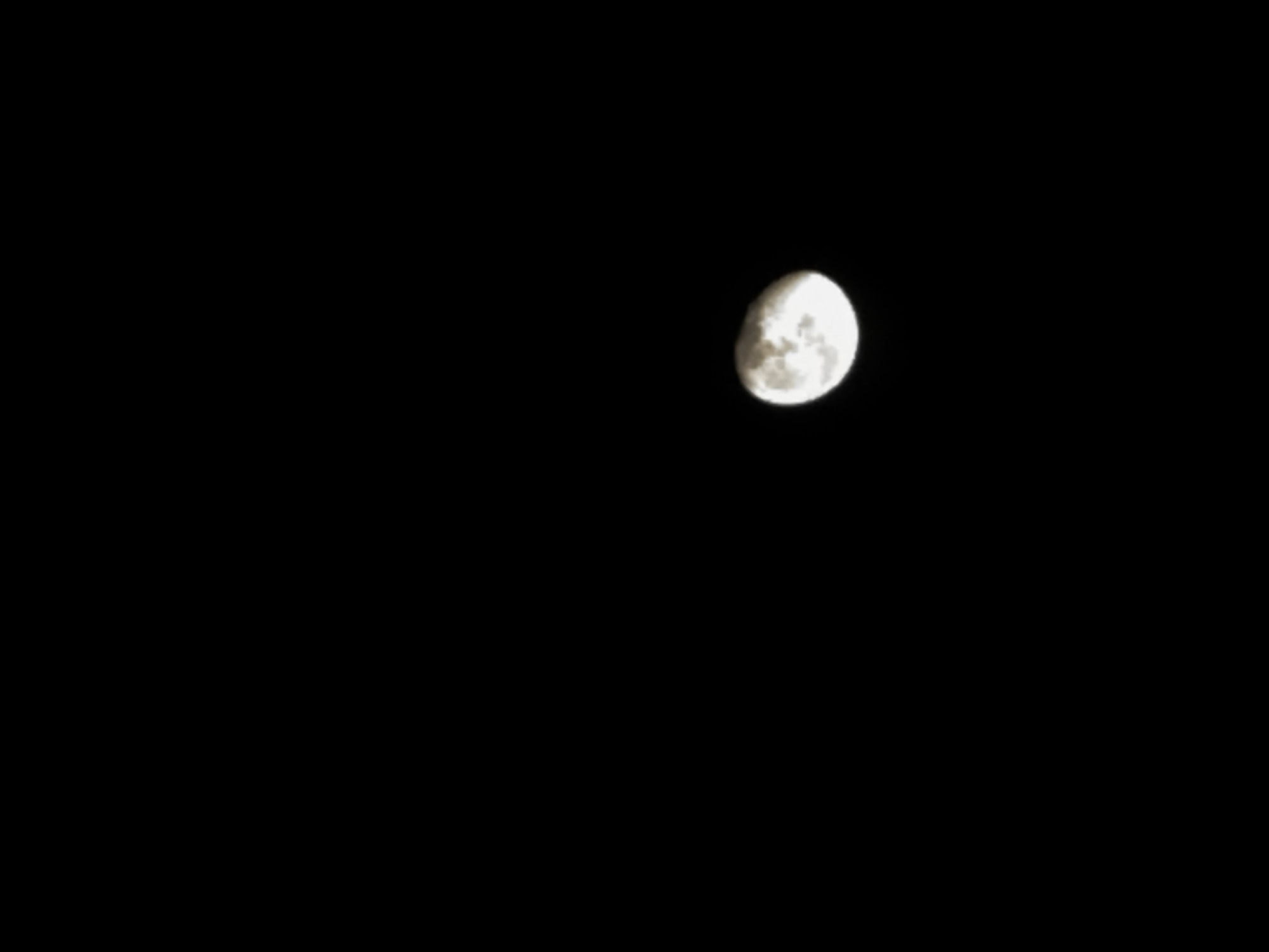 A close-up, night-time photograph captures the moon in the upper right quadrant of the frame. The moon appears as a bright, glowing orb, slightly more than half-illuminated, with visible craters and surface textures. The surrounding background is entirely black, indicating a very dark sky. There are no other objects or subjects visible in the image. The location is Unknown, Unknown.