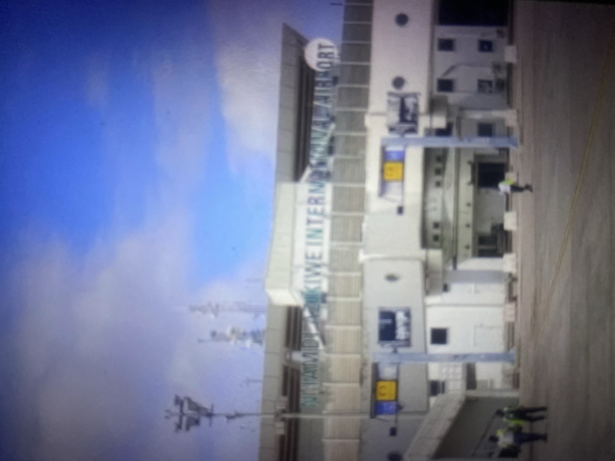 The image shows a blurry exterior view of an airport terminal building. Above the terminal, the words "NTAMIDE KINWE INTERNATIONAL AIRPORT" are visible in large green lettering, though partially obscured and out of focus. The building is primarily white with a grey, sloped roof. There are several windows and circular elements on the facade. A ramp or walkway leads to the left side of the building. In front of the terminal, there is a paved area, presumably an apron or tarmac. Two figures, appearing to be men, are walking on this paved area. One is wearing a light-colored shirt and dark pants, and the other is wearing a dark uniform with a yellow vest. The sky above is blue with some scattered white clouds. The overall image quality is low, with significant blur and pixelation.