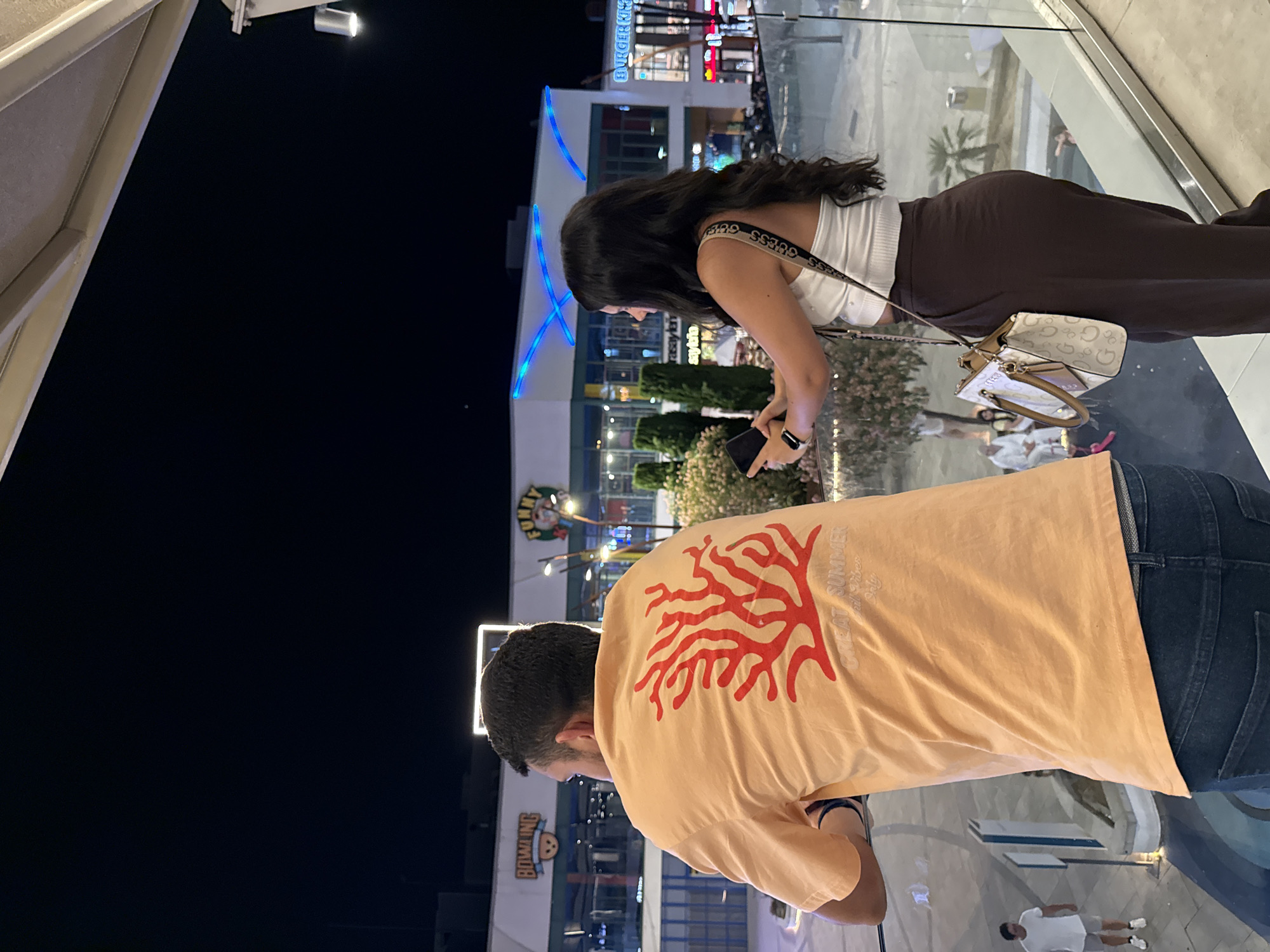 This media file shows two people, a woman and a man, in an outdoor urban setting at night. The woman, in the foreground on the right, is wearing a white crop top and dark brown pants. She has dark hair and is looking down at her phone, which she holds in her right hand. She also has a purse with a patterned strap and a light-colored bag. The man, in the foreground on the left, is wearing a peach-colored t-shirt with a red coral-like graphic and dark jeans. He is also looking downwards.
In the background, there is a multi-story building with illuminated signage. One sign reads "BURGER KING" in blue letters. Another sign says "BOWLING" with a cartoon character. There are also trees and other people visible in the distance, as well as lights reflecting on a surface, suggesting a reflective floor or water feature. The scene appears to be an entertainment or shopping area. The lighting suggests it is nighttime. The overall atmosphere is casual and urban. The text visible includes "BURGER KING", "BOWLING", and "GREAT SUMMER".