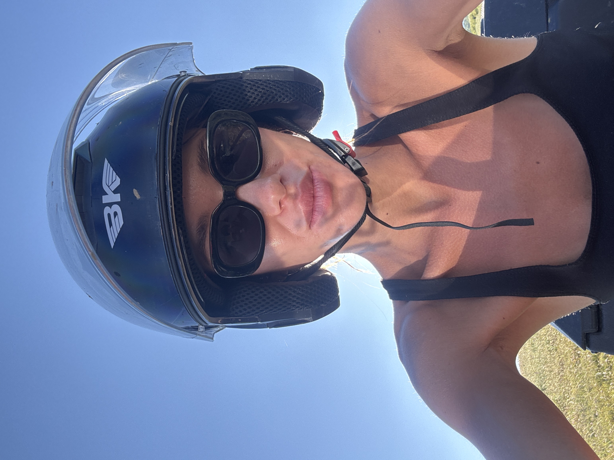 A woman wearing a black helmet, black sunglasses, and a black tank top is shown in a close-up, low-angle selfie. The sun is shining brightly, creating highlights on her skin and casting shadows. Her hair is visible beneath the helmet, and it appears to be light brown. The background is a clear blue sky with a hint of grass and possibly distant trees. The overall impression is that she is outdoors, possibly on a sunny day, and appears to be riding a vehicle, given the helmet. There are no identifiable landmarks or clues to suggest the location is Tbilisi, Georgia.