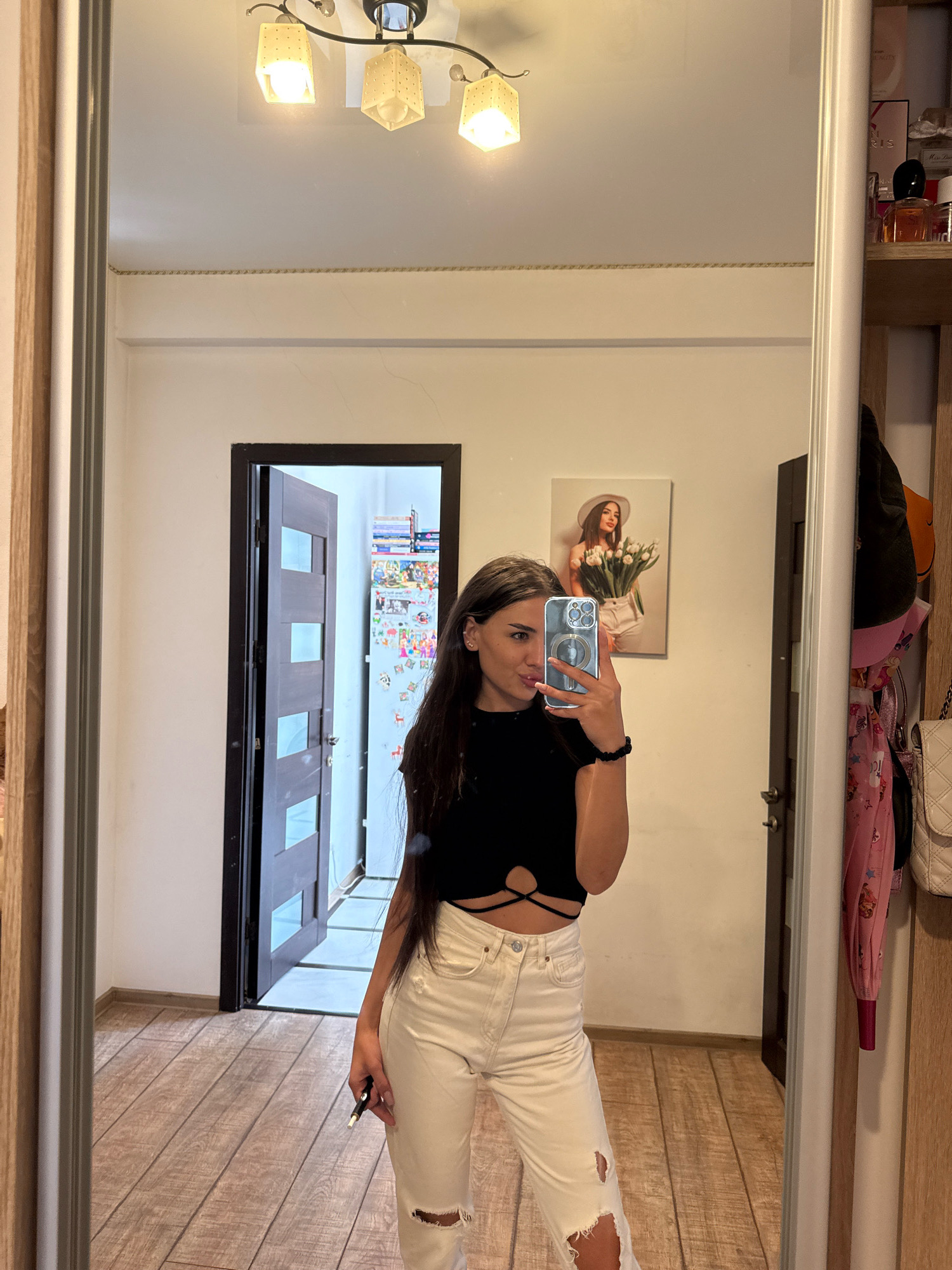 The image shows a young woman taking a mirror selfie in what appears to be an interior hallway. She is wearing a black crop top with a cutout design and light-colored ripped jeans. She is holding a phone in her right hand and a pen in her left. In the background, a door is open, revealing more of the interior, including shelves with items and what looks like a refrigerator covered in pictures and notes. A framed picture of a woman holding flowers hangs on the wall to the left of the door. The floor is made of wooden planks. The lighting suggests it is daytime. No specific landmarks or indicators place the scene within Chișinău, Moldova.