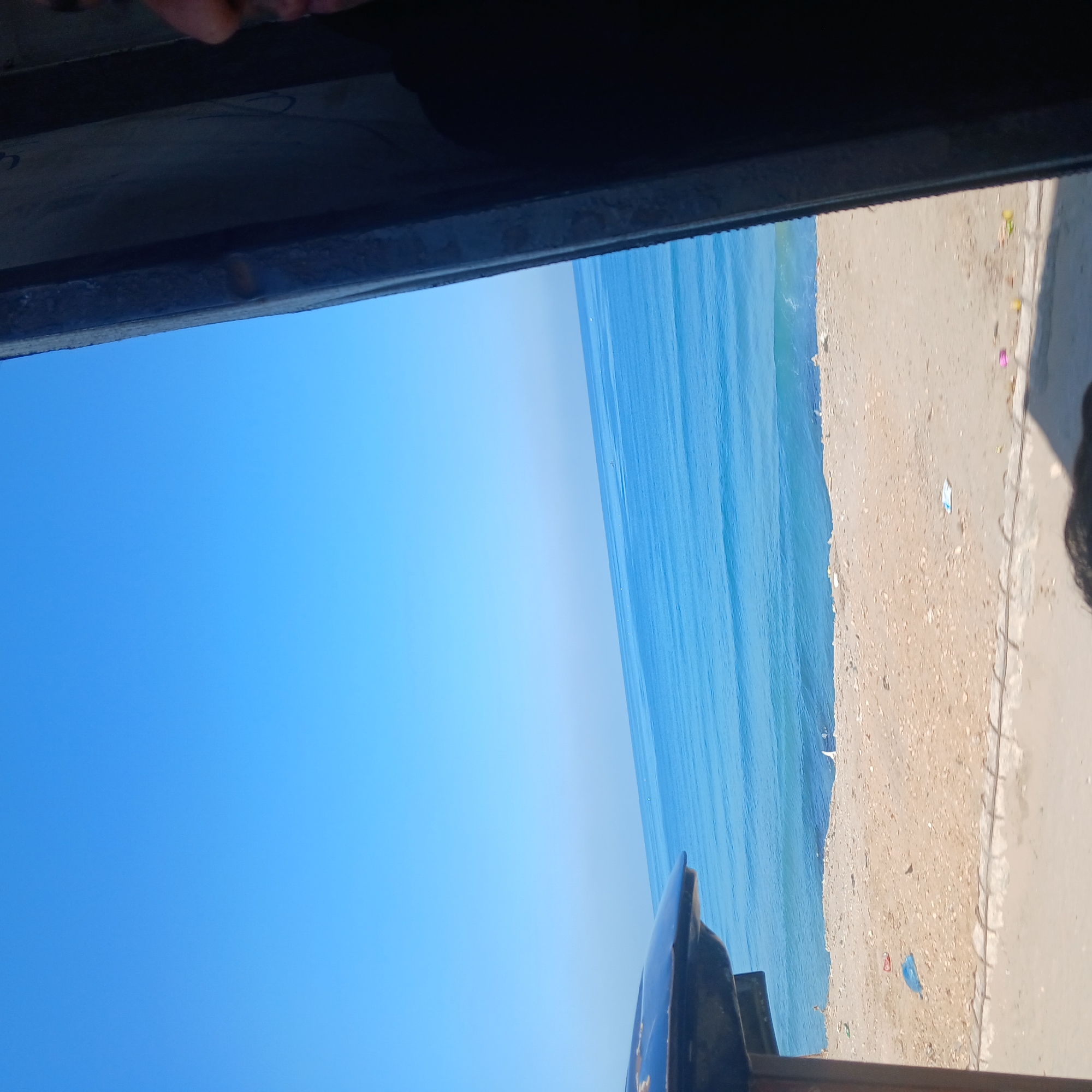 The image, originally captured sideways, displays a bright coastal vista when viewed with the sky at the top.
**Subjects:** The scene features a vast expanse of clear blue sky, a wide blue ocean, and a sandy beach. In the immediate foreground (at the bottom of the correctly oriented image), there is a dark, solid horizontal structure, likely a railing or the edge of a window/balcony from which the photo was taken. Dark, blurry, and out-of-focus shapes in the bottom-left corner suggest parts of people looking out. The beach shows small, colorful pieces of litter, including red, blue, and yellow items.
**Activity or event taking place:** The image depicts a view from a vantage point, suggesting someone is observing the coastal landscape.
**Notable details:** The clear, bright blue sky indicates sunny weather during the day, likely mid-day as there are no prominent shadows. The ocean has visible ripples on its surface. The beach transitions into what appears to be a paved or concrete edge before the foreground structure. The wide perspective captures a significant portion of the sea and sky.
**Visible text:** There is no discernible text in the image.
