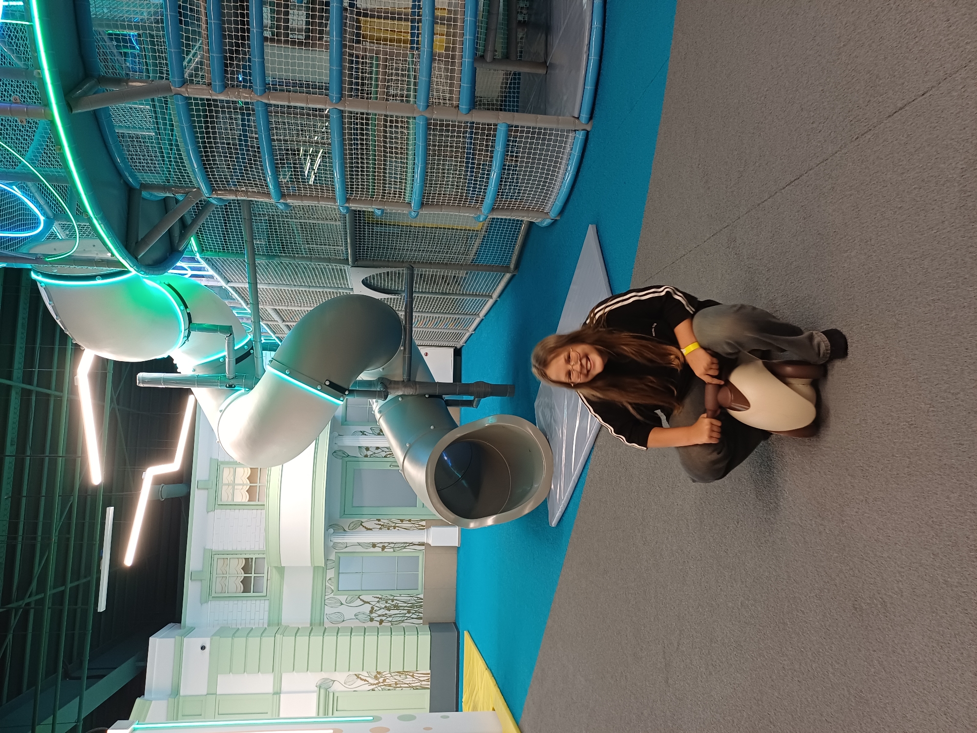 A young female with long brown hair and glasses is sitting on a toy, in a brightly lit indoor play area. She is wearing a black long-sleeved shirt with white stripes on the sleeves, and dark pants. She is smiling. The play area features a large, multi-level structure with slides, netting, and blue and green neon lights. The floor is covered in blue and gray carpeting, with a yellow mat on the left. The background includes a decorative facade resembling a building with windows and columns. The lighting is artificial and bright, creating a vibrant atmosphere. The image is taken from a medium distance, capturing the subject and a significant portion of the play structure.
