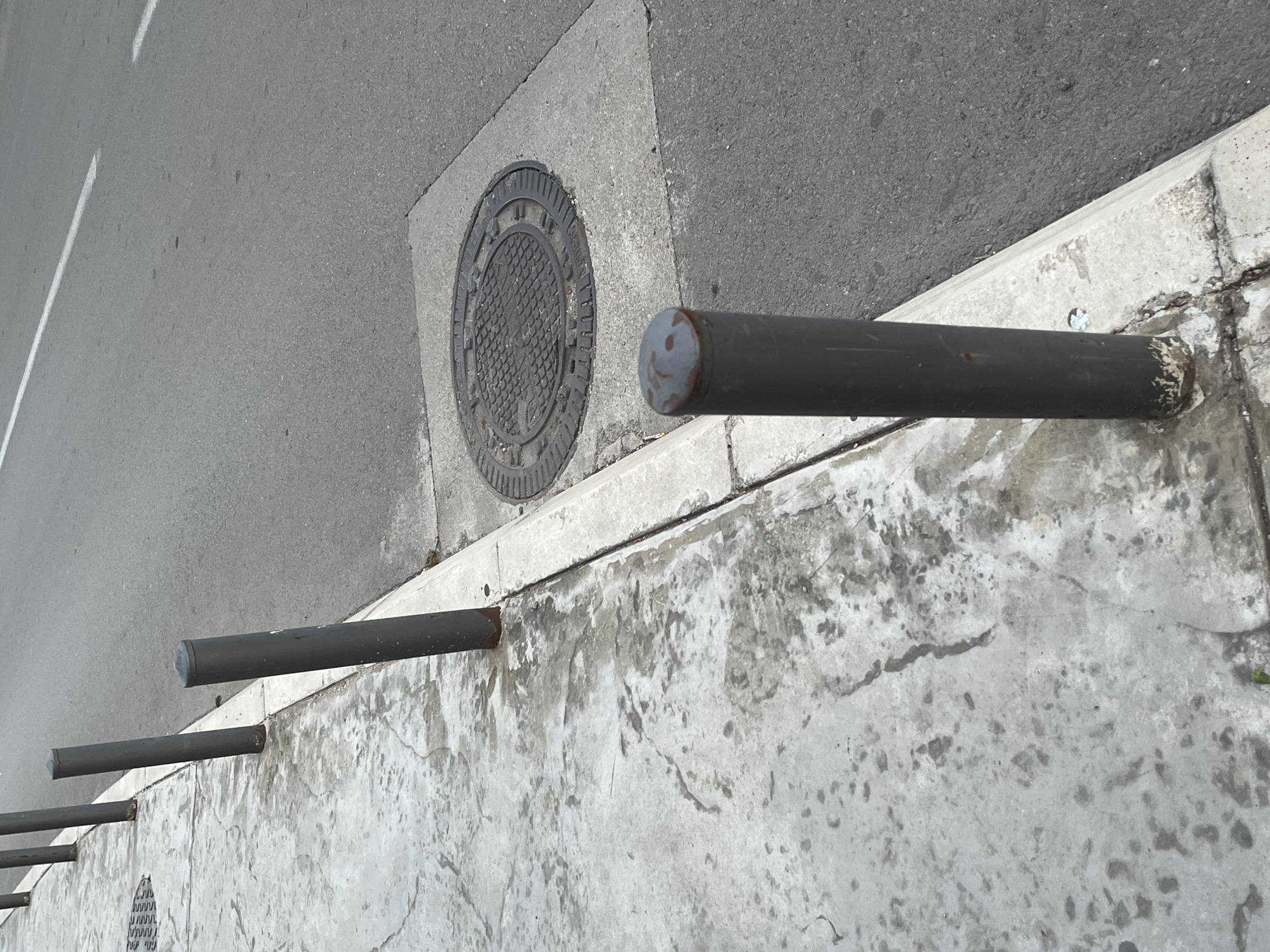 The image shows a row of dark grey bollards along a sidewalk in Budva, Montenegro. The sidewalk is made of concrete and appears somewhat worn and stained. To the right of the sidewalk is a paved road with a white lane marker. In the road, a manhole cover is visible, set within a concrete frame. The scene is outdoors and the lighting suggests it is daytime. No people are present in the image.
