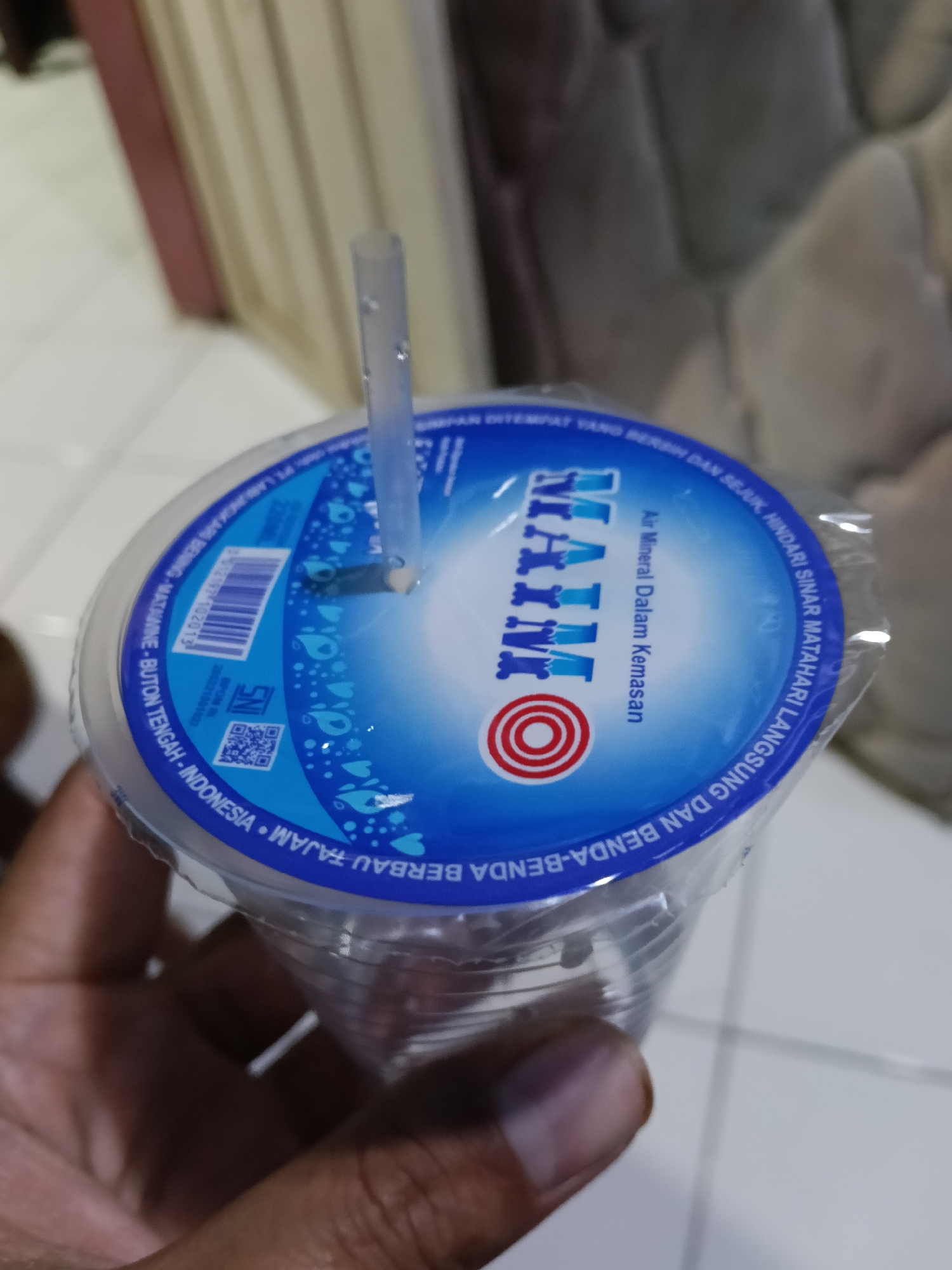 A clear plastic cup filled with water is held in a person's left hand. The cup has a blue and white lid with text in Indonesian. A clear plastic straw extends from the lid. The background is blurred and shows a patterned surface and a vertical structure. The image is a close-up shot, with the cup and hand dominating the frame. The scene appears to be indoors.