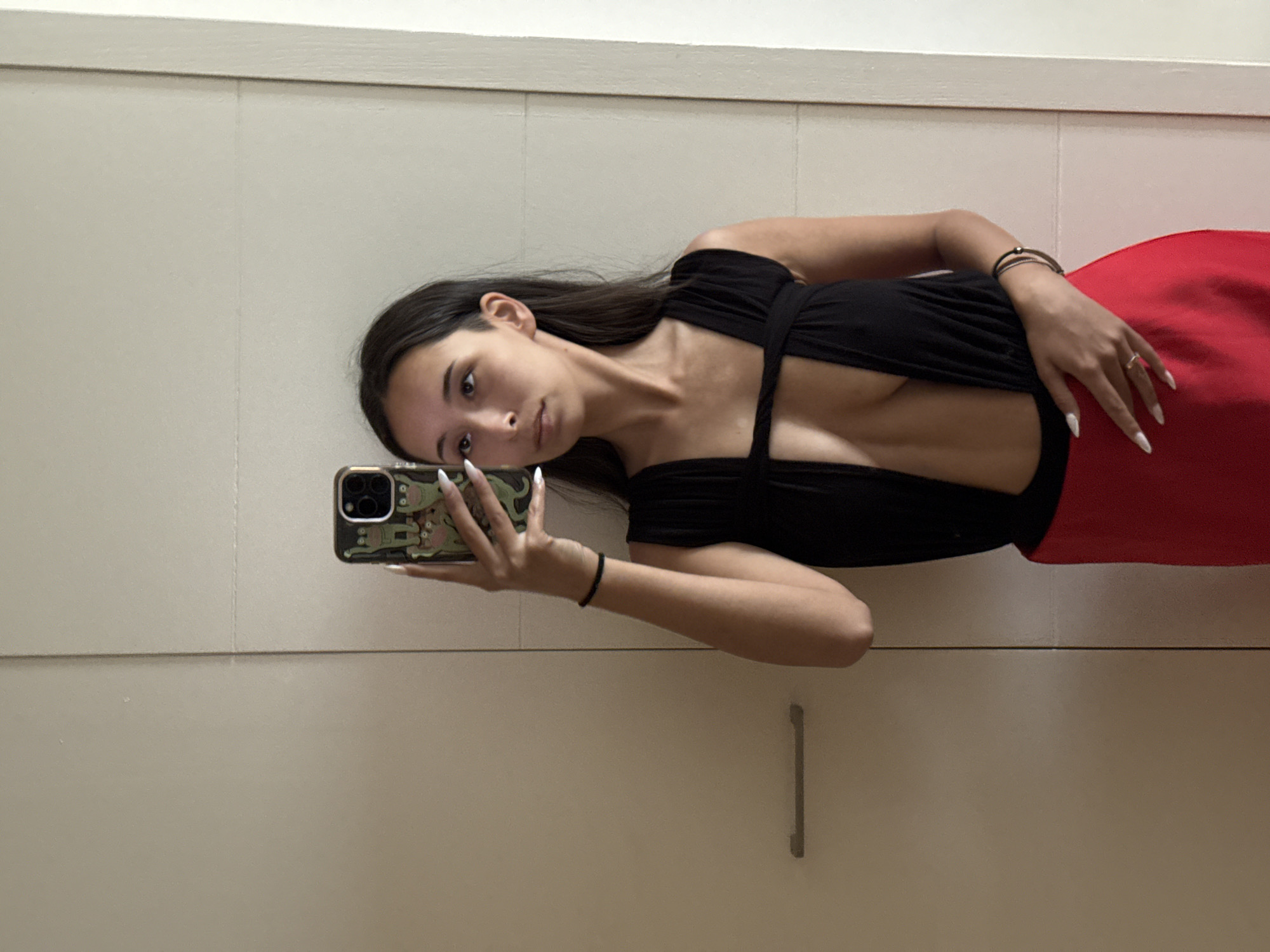 A young woman with dark hair and brown eyes is taking a selfie in a mirror. She is wearing a black top with a deep V-neck and a red skirt. She has on white acrylic nails and is wearing a ring on her left ring finger, and two bracelets on her left wrist. She is holding her phone, which has a frog-themed case. The background appears to be a closet or a large cabinet. The location is not specified in the image or context.