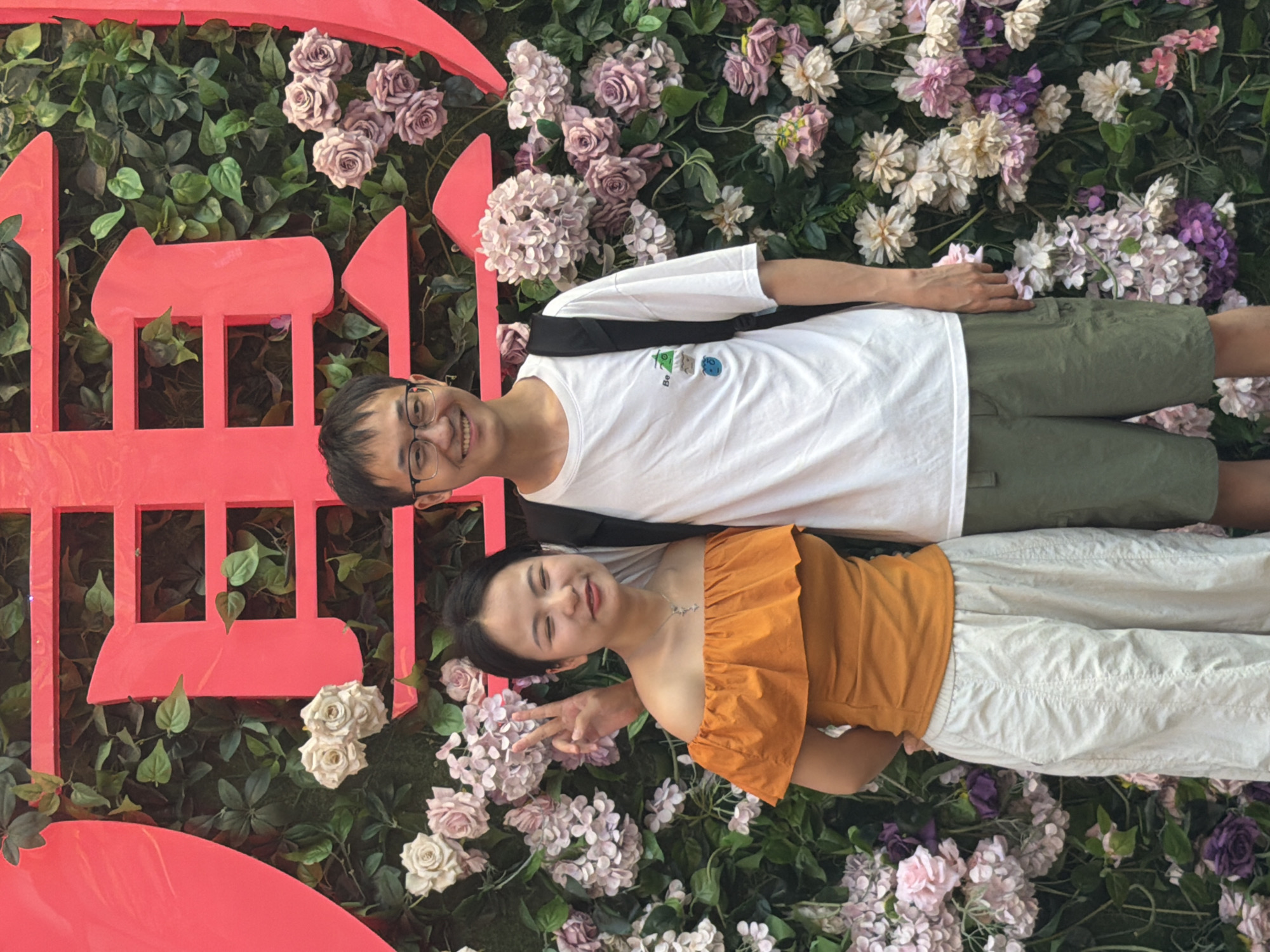 The image shows a young Asian couple posing for a photo in front of a floral wall display with large red characters, possibly in Yuzhong District, China. The woman is on the left, wearing an off-the-shoulder orange top and light-colored pants, and is making a peace sign with her fingers. The man is on the right, wearing a white t-shirt with a graphic print, khaki shorts, and glasses, with a backpack slung over his shoulder. The floral wall features a mix of green foliage and various shades of pink, purple, and white artificial flowers, resembling hydrangeas and roses. The red characters appear to be Chinese calligraphy, large and prominently displayed behind the couple. The lighting suggests it is daytime, possibly outdoors or in a well-lit indoor space.