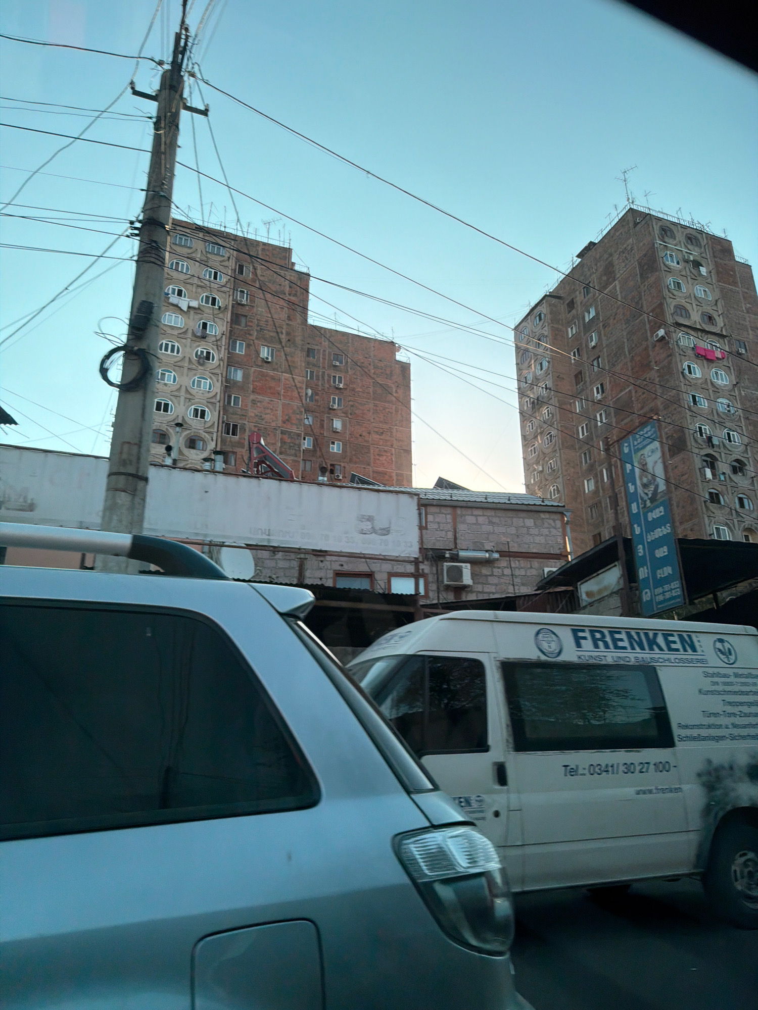 The image depicts a street scene in Yerevan, Armenia, with prominent Soviet-era apartment buildings visible in the background. In the foreground, a silver car is partially visible on the left, and a white van with "FRENKEN KUNST UND BAUSCHLOSSEREI" written on its side is on the right. The van appears to be a service vehicle, possibly for a metalworking or construction business. Overhead, a complex network of power lines crisscrosses the sky, typical of urban infrastructure. Laundry is seen hanging from some of the apartment windows. The overall atmosphere suggests a typical urban environment in Yerevan, characterized by older architecture and active street life.