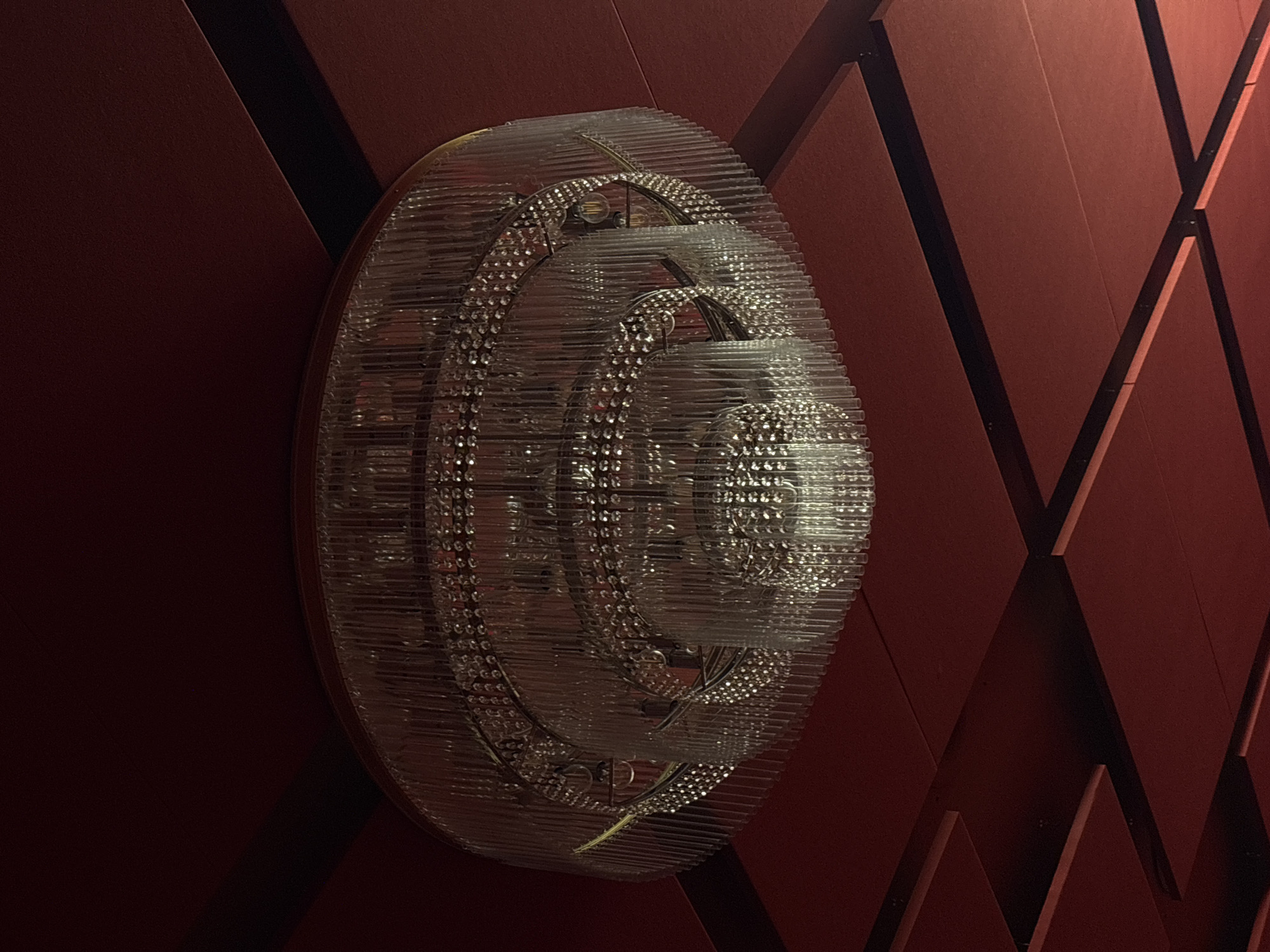The image depicts a decorative wall light fixture, possibly made of glass or acrylic tubes arranged in concentric circles, against a geometrically patterned wall. The wall features reddish-brown panels with black outlining, arranged in a way that creates a series of diamond and triangular shapes. The lighting suggests an indoor environment, and the fixture is likely for aesthetic purposes. There are no people visible in the frame, and the focus is on the interplay between the light fixture and the wall's design. The location, Minsk, Belarus, is not discernible from the visual content of the image.