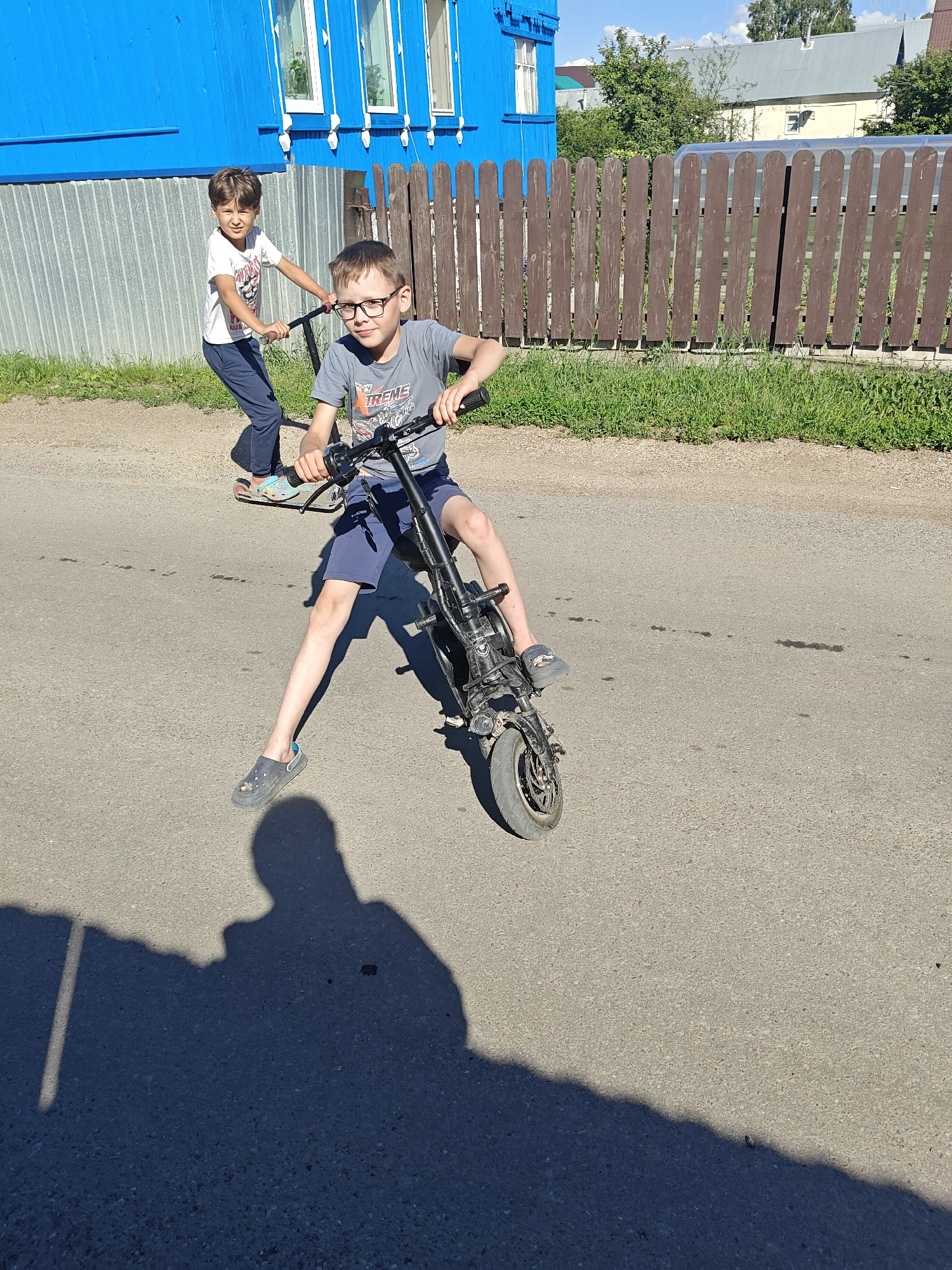 Two boys are riding scooters on an asphalt road in front of a blue wooden house and a brown wooden fence. The boy in the foreground is wearing glasses, a gray t-shirt, and blue shorts. He is on a black electric scooter. The boy in the background is wearing a white t-shirt and blue pants, and is on a kick scooter. The scene appears to be in a rural or suburban area, with green grass and trees visible. The sun is shining, casting shadows on the road.