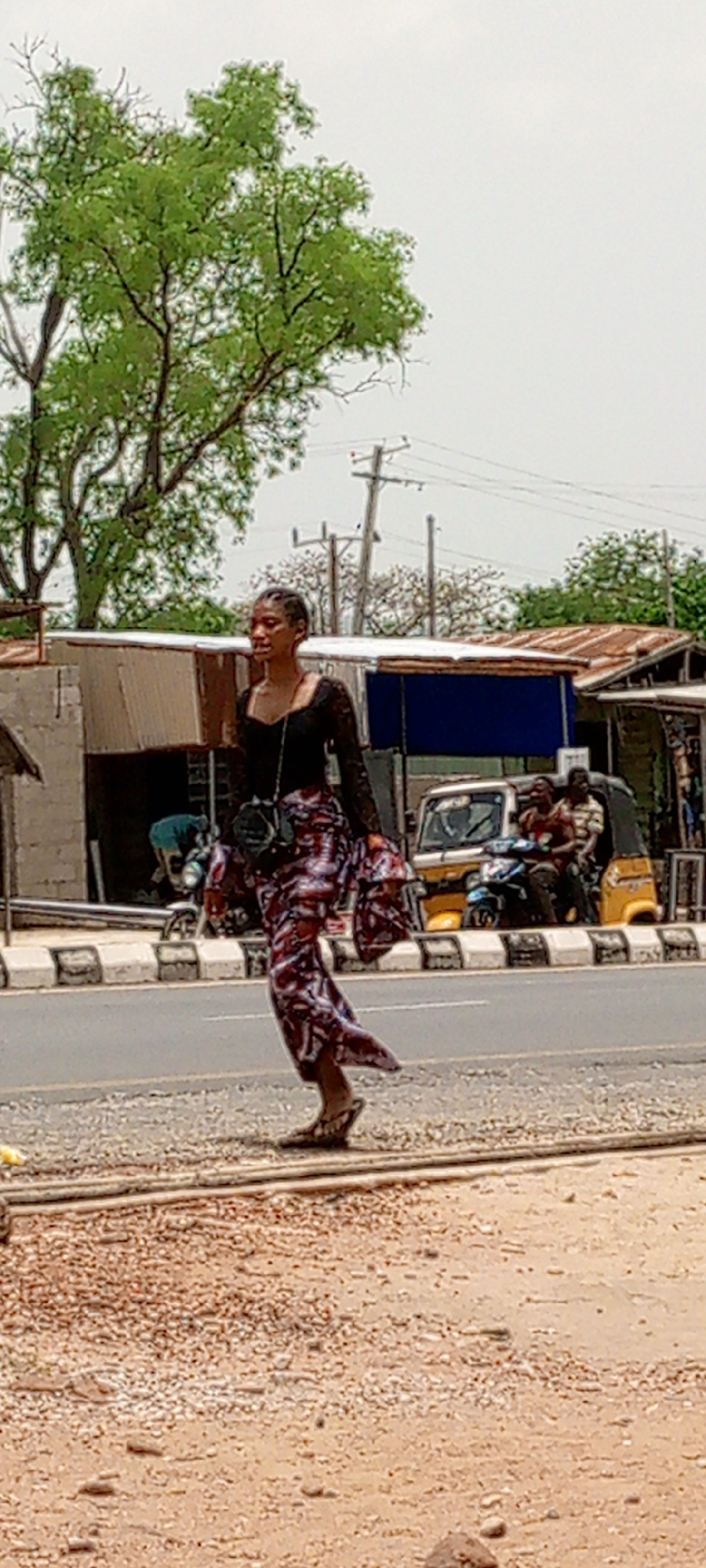 A woman in a long patterned dress and black top walks near a road in Adankolo, Nigeria. She carries a small black bag. Behind her, a yellow vehicle with passengers is visible near buildings with corrugated metal roofs. A large tree is to the left, and power lines are in the background.