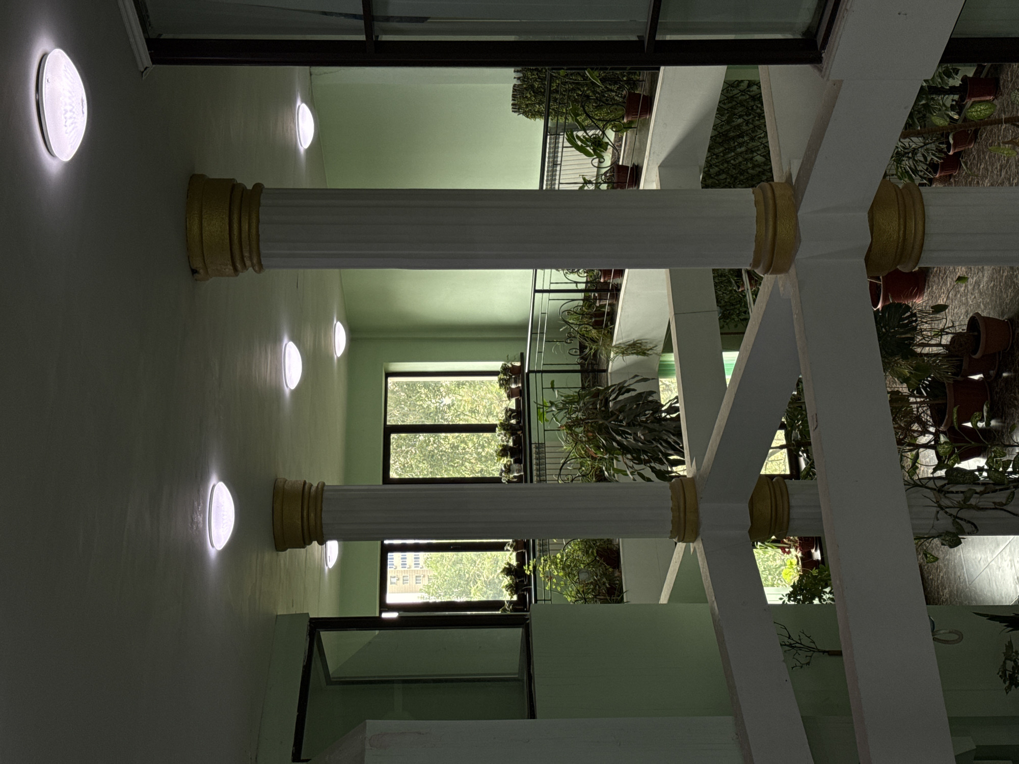 The image shows an interior view of a building with a central atrium. The walls are painted a pale green, and the ceiling is also a light color with several circular, flush-mount light fixtures. Two prominent, fluted white columns with gold trim support the ceiling. Balconies or walkways, also with white railings and some with decorative metalwork, are visible on different levels.
Numerous potted plants are strategically placed throughout the space, along the balconies and on the floor. Some plants are small and neat, while others are larger and more sprawling. Through windows, glimpses of trees and an urban building are visible, suggesting the location is in a city. The flooring appears to be tiled. The overall impression is of a well-maintained, plant-filled indoor space, possibly a common area in a residential building or a commercial space designed with an emphasis on natural elements.
Based on the architectural style and the use of such a plant-filled atrium, this could be a common area within a residential complex or a public building in Almaty, Kazakhstan. Such interior courtyards are sometimes found in modern buildings in the city.
