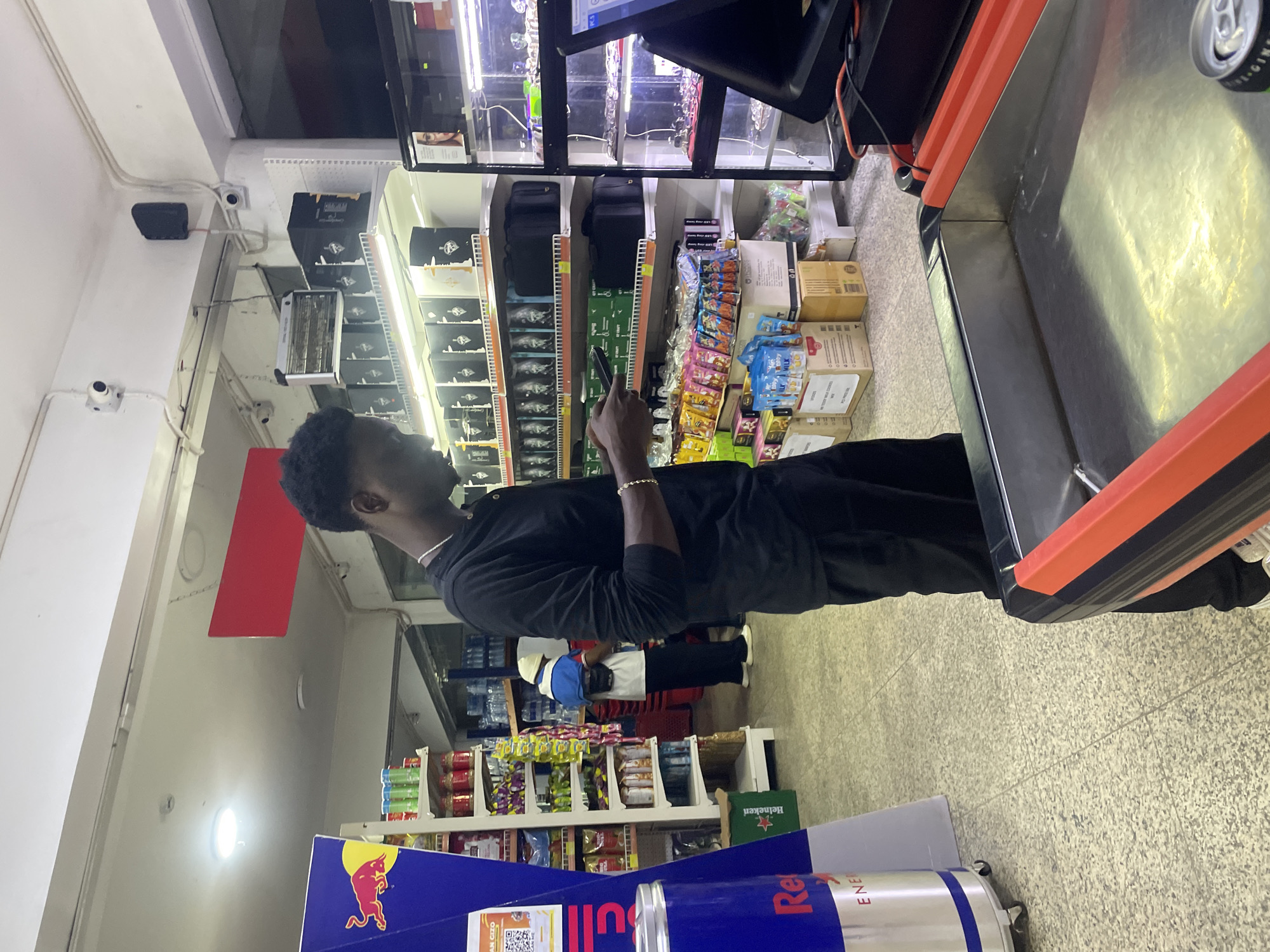 This image depicts a retail environment, likely a convenience store or small supermarket, located in Galadimawa Village, Nigeria. In the foreground, a young Black male is standing, facing a checkout counter, and is looking down at his mobile phone. He is wearing a black long-sleeved shirt and black pants, with a silver necklace and bracelet. To the left of the frame, a tall, cylindrical Red Bull display cooler is visible. Behind the main subject, shelves are stocked with various products, including drinks, packaged snacks, and some boxed items. Further in the background, another individual wearing a light-colored hat and a blue and white top is seen browsing the shelves. The store appears well-lit, with overhead fluorescent lights and some visible security cameras and fixtures on the ceiling. The flooring is a speckled tile. The overall atmosphere suggests a typical day of commerce in a local Nigerian community.