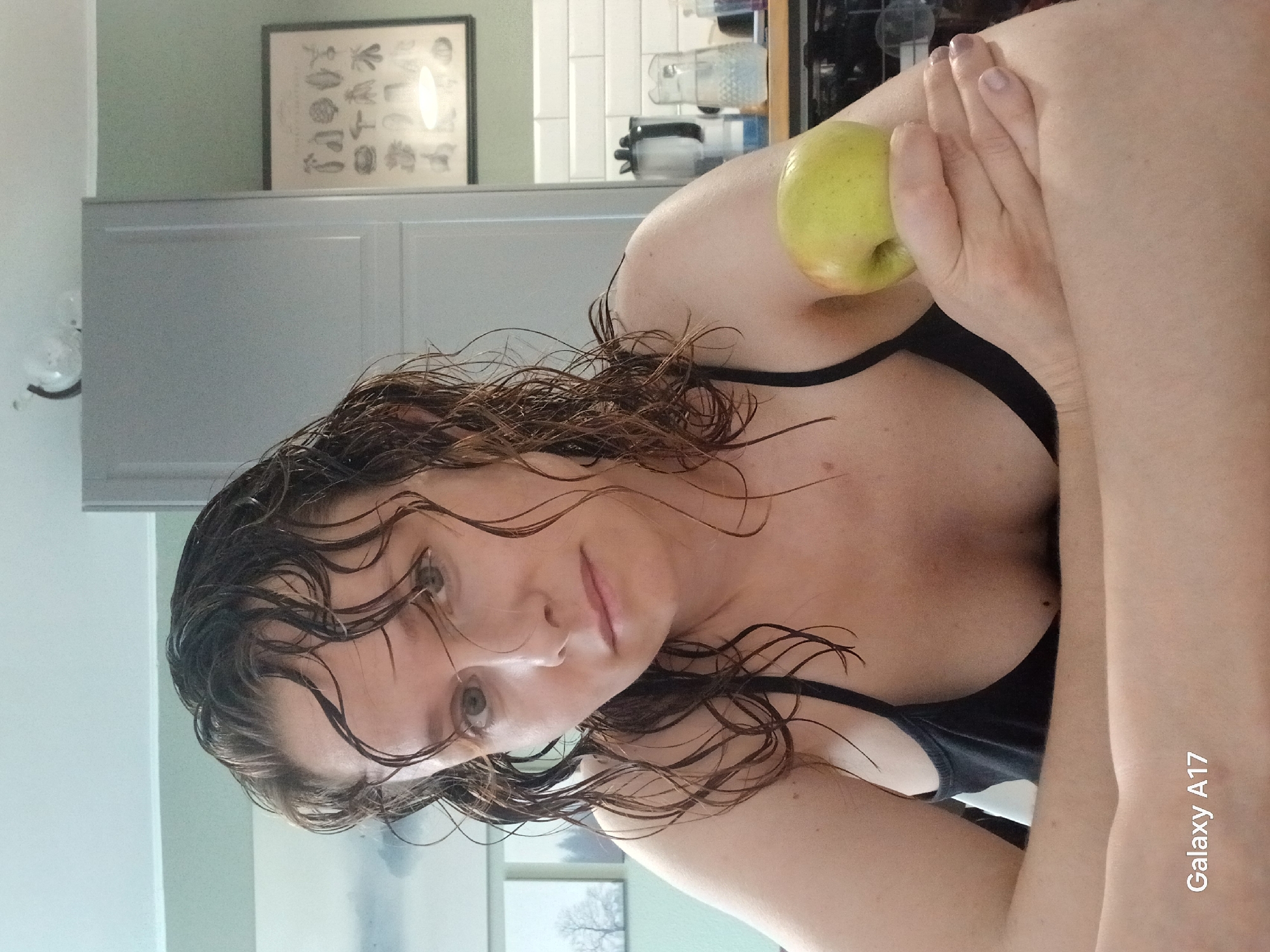 A woman with wet, wavy brown hair poses indoors, holding a green apple against her right shoulder and arm. She wears a black top and faces the camera. The background shows a kitchen with white subway tiles, kitchen appliances, and a framed botanical print on the wall. The image is oriented vertically. The location in the city of Yekaterinburg, Russia is unknown.