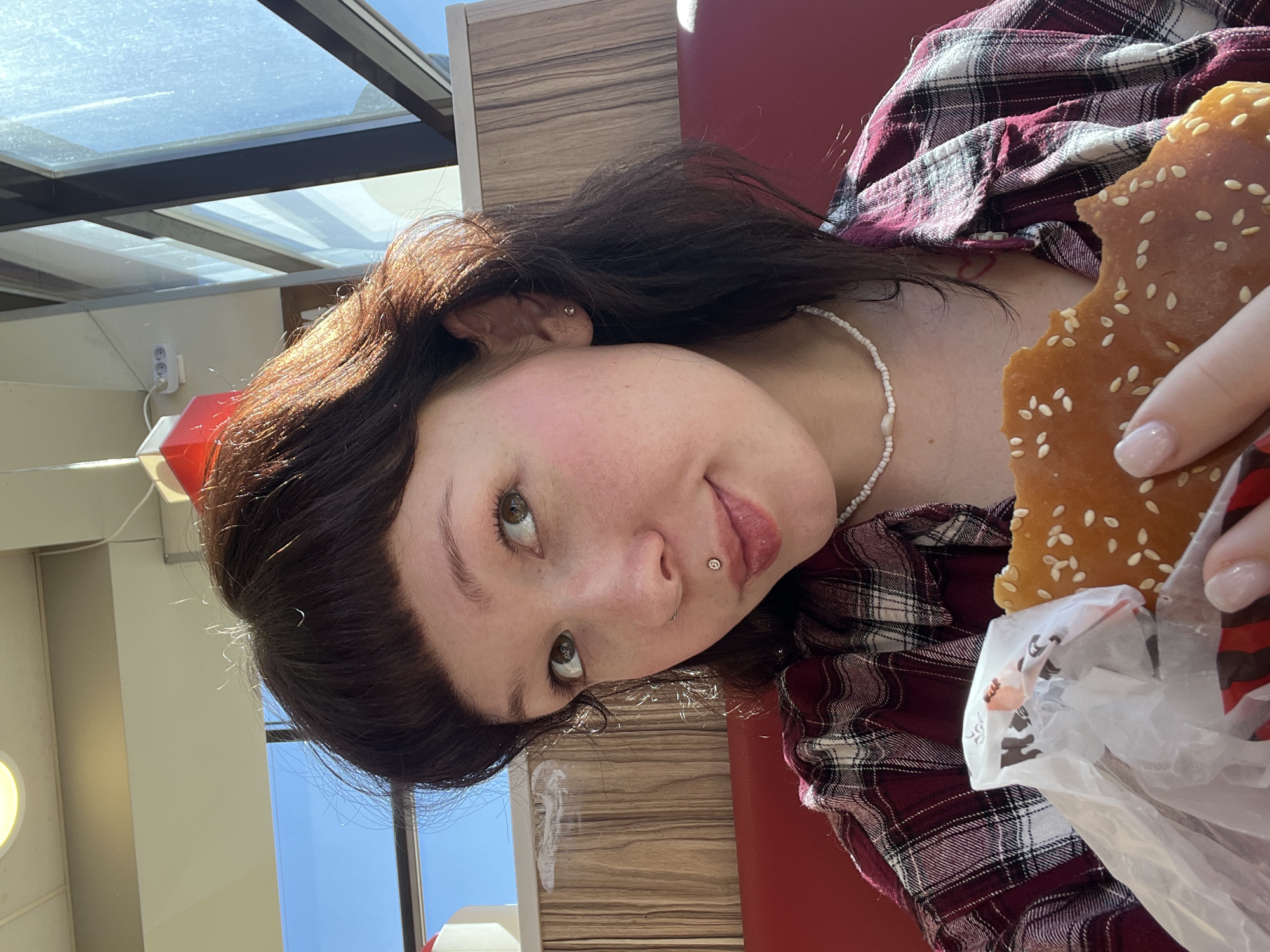 The image displays a young person indoors, likely within a fast-food restaurant or cafe in Izhevsk, Russia, during a bright, sunny daytime. The subject, a person with dark brown hair styled with bangs, is captured at an angle, looking directly at the viewer with a neutral expression. They have a fair complexion with rosy cheeks, and visible piercings include a shiny stud in their philtrum and a simple stud earring in their ear.
They are wearing a red and white plaid shirt and a white beaded necklace. In their hand, they hold a large, partially eaten bun topped with sesame seeds, characteristic of a burger bun. The bun is wrapped in a white paper with indistinct red and black branding elements. Their fingernails are painted with a light, neutral polish.
The background features bright natural light filtering through a large glass ceiling or skylight structure, confirming the daytime setting. A red upholstered seat or booth and a wooden-patterned wall or partition are visible, contributing to the restaurant ambiance. An electrical outlet with three sockets and a red object are also present in the upper left background.