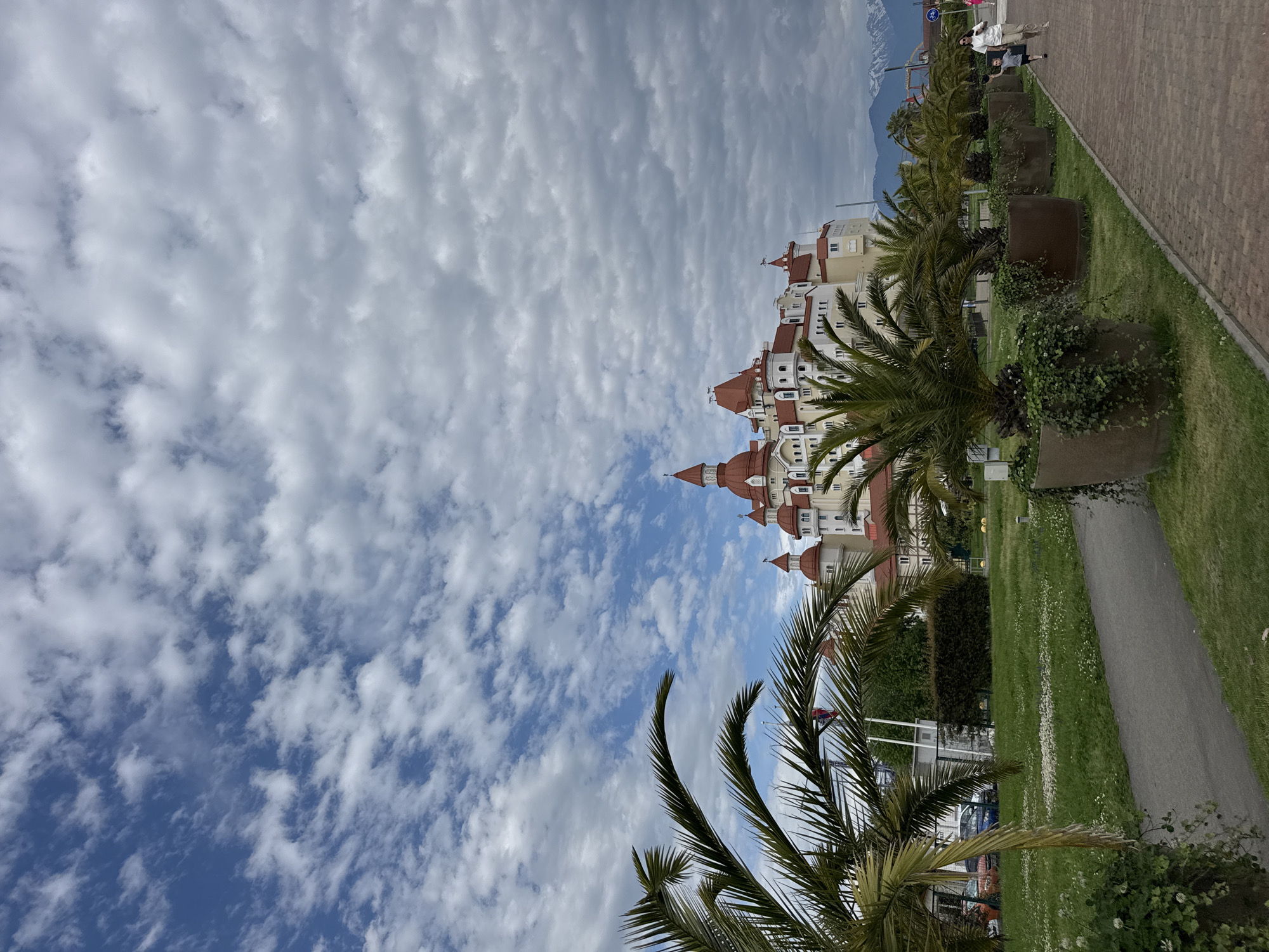 The image depicts a building with red-tiled roofs and white walls, possibly a hotel or resort, in Sirius, Russia. The building is situated along a grassy area with palm trees and paved walkways. The sky is filled with white, puffy clouds. In the distance, there are mountains. There are also some people walking along the sidewalk.