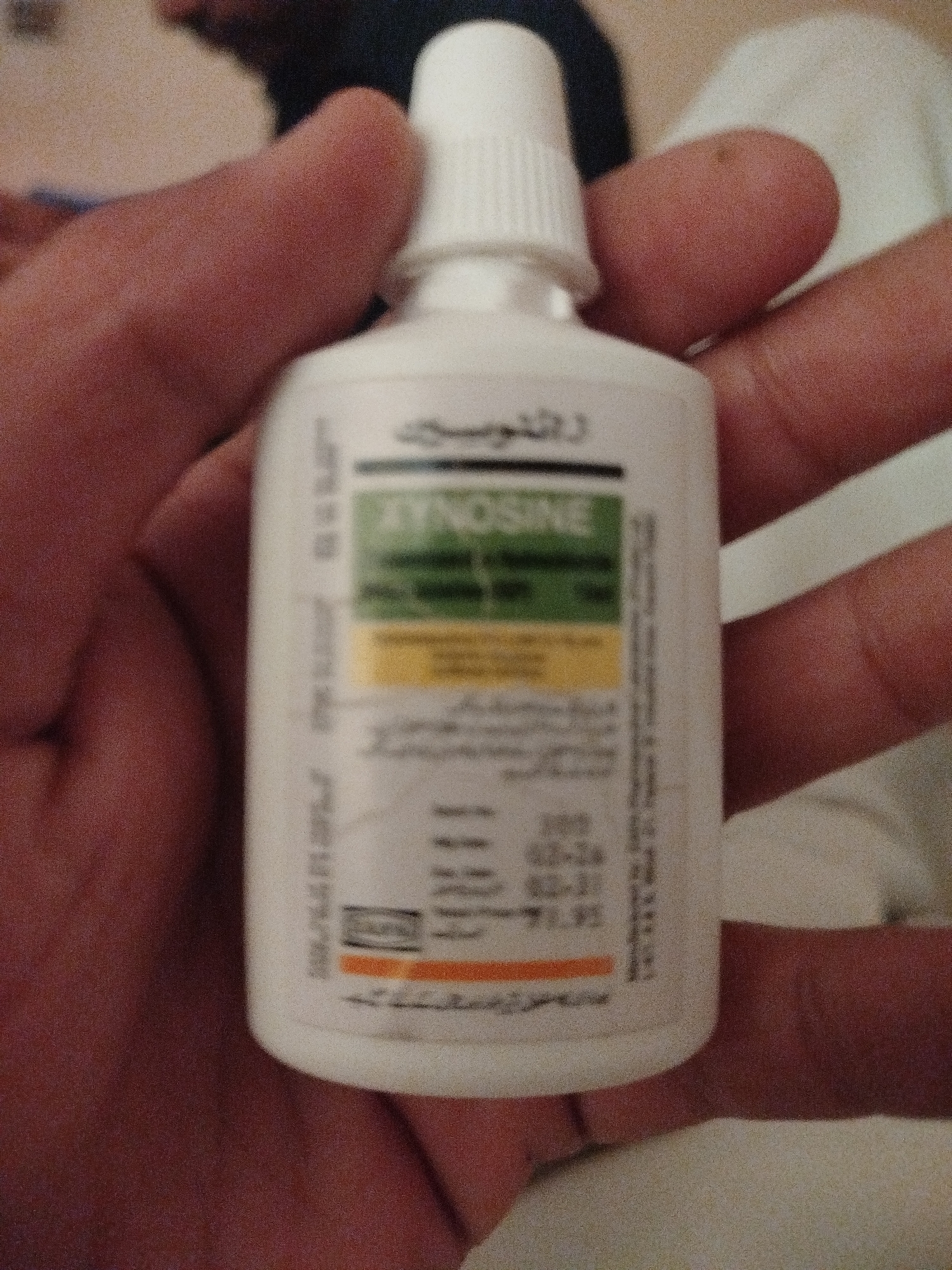A person's hand holds a white plastic bottle with a white cap. The bottle has a label with text and colored sections. The label is mostly in Arabic. A green rectangular area on the label contains the text "ZYNOISINE" in white letters. Below that, a yellow rectangular area has some Arabic text. Further down, there is a barcode and more Arabic text, along with what appears to be a date and lot number: "200", "02-26", and "02-31". The image is blurry and the background is out of focus, showing hints of fabric and a dark color, possibly clothing or a blanket. The image location is not determinable from the visual information provided.