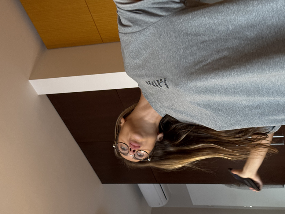 An indoor image depicts a female subject with long, multi-toned brown hair featuring blonde highlights. She wears round, dark-rimmed glasses and a grey short-sleeved t-shirt with black text on the left chest, partially legible as "If I'm late, I didn't want to come,". Her head is tilted sharply upward and to her right, with a pouting expression. Her left hand holds a dark-colored smartphone, visible near her lower body. The background consists of a light-colored ceiling, a dark brown wall on the left with a white rectangular air conditioning unit mounted high, a white vertical pillar or wall section in the center-right, and a light yellowish-brown wall or cabinetry on the far right. No identifying architectural or geographical markers are present to determine a specific location within Warsaw, Poland.