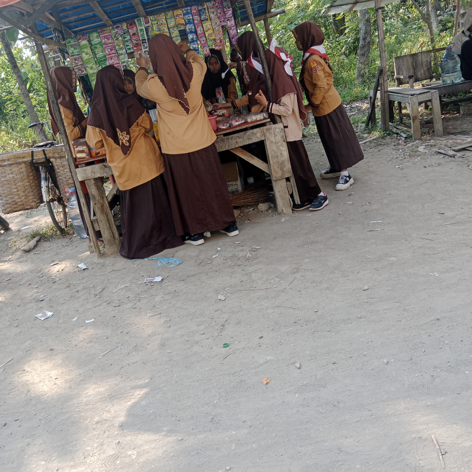 In Tegowanu Kulon, Indonesia, a group of schoolchildren, likely students from a local school given their uniforms, are gathered around a makeshift roadside stall. The stall is covered with a blue tarp supported by wooden poles and displays various packaged snacks and drinks. The children are wearing tan-colored uniforms with brown hijabs, and some have scout-like red and white neckerchiefs. They appear to be in the process of purchasing or browsing items from the stall. One girl in the center is looking towards the stall owner. The ground is unpaved, covered with dirt and scattered debris. To the left, a bicycle with a woven basket is parked, and a large bottle of water sits near the stall. In the background, lush green foliage and trees are visible, suggesting a rural or semi-rural setting. The overall scene depicts a common everyday interaction at a local village store.