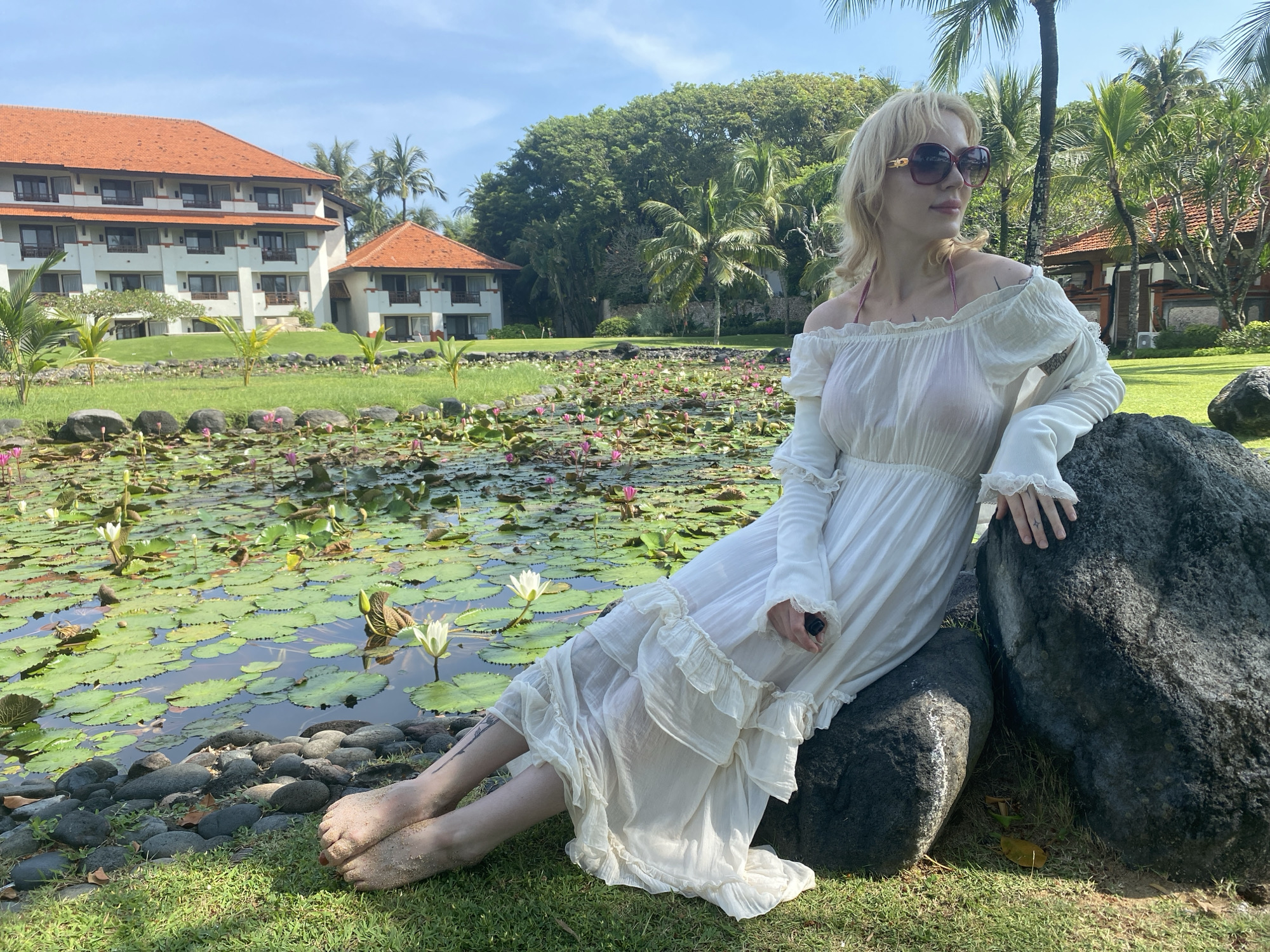 A person is sitting on a rock in an outdoor setting near a pond filled with lily pads and lotus flowers. The individual is wearing a white, off-the-shoulder long-sleeved dress and large sunglasses. The person's bare feet are visible, and they appear to be sandy.
In the background, there is a building with a terracotta-tiled roof, surrounded by lush green trees and palm trees. This appears to be a resort setting. The image was taken in Nusa Dua, Indonesia.