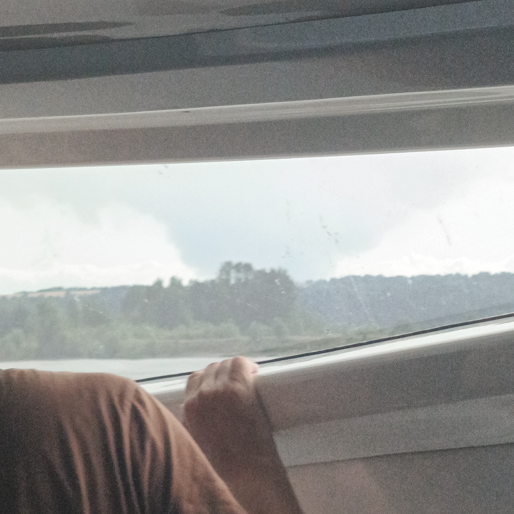 A close-up shot captures a person's arm and hand resting on a window ledge, looking out at a landscape of trees and a cloudy sky. The person is wearing a brown shirt, and their hand is gripping the white window frame. The view outside is slightly blurred, with a hint of water visible in the foreground. The overall impression is one of a journey or a moment of quiet observation.