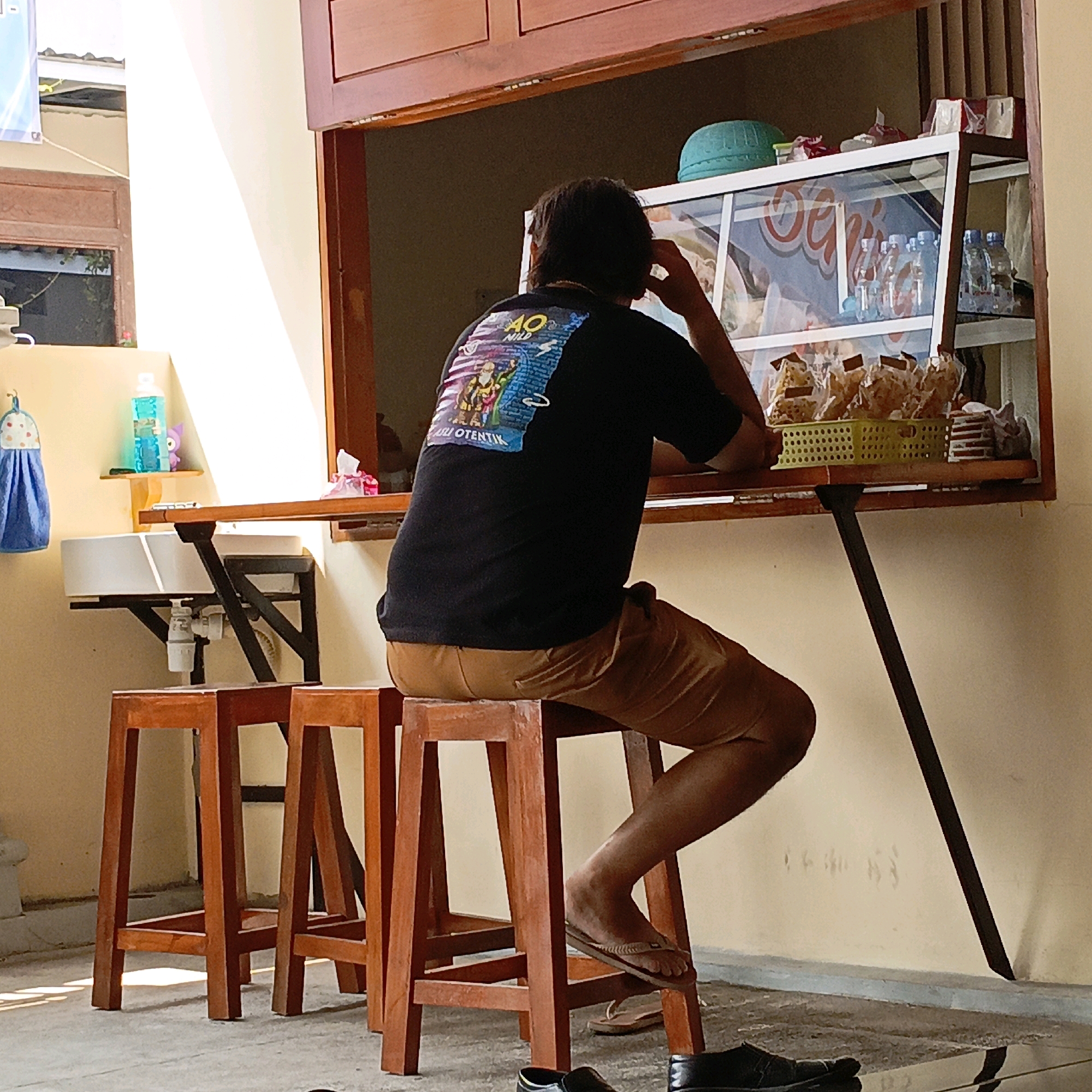 In Salatiga, Indonesia, a person is seated on a wooden stool at a counter, facing away from the camera. The individual wears a black t-shirt with a colorful graphic and tan shorts. The counter is in front of a display case filled with bottled drinks and packaged snacks. To the left of the counter, a white sink is mounted, with a blue towel hanging nearby. The scene appears to be an outdoor food or drink stall, with sunlight casting shadows on the ground. The environment is simple and functional.