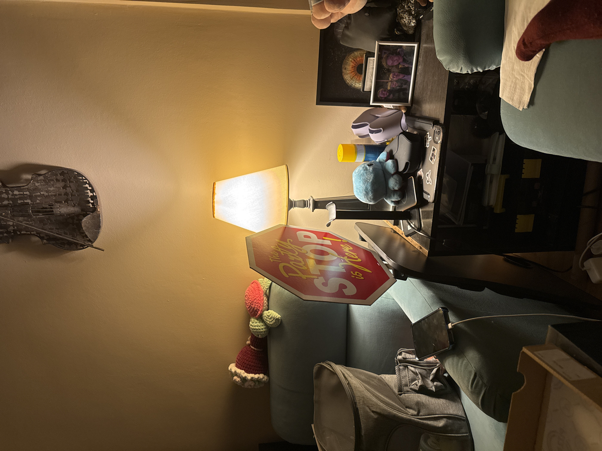 A cluttered interior scene features a lamp illuminating a seating area. A wall decoration resembling a violin hangs on a tan wall. A stop sign parody decoration leans on a light-blue couch where a yarn doll resides. A gray bag rests on the couch, and a black desk is visible with items on top, including a picture frame, headphones, and a blue stuffed animal. The location is not discernible from the provided image.