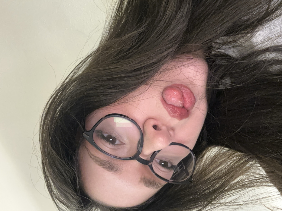 An upturned face is framed by dark hair. A young woman wears round black glasses. Her expression is playful, with her tongue sticking out and pressed against her upper lip. The camera angle gives a distorted perspective. The background is blurred, suggesting a close-up shot with a shallow depth of field.