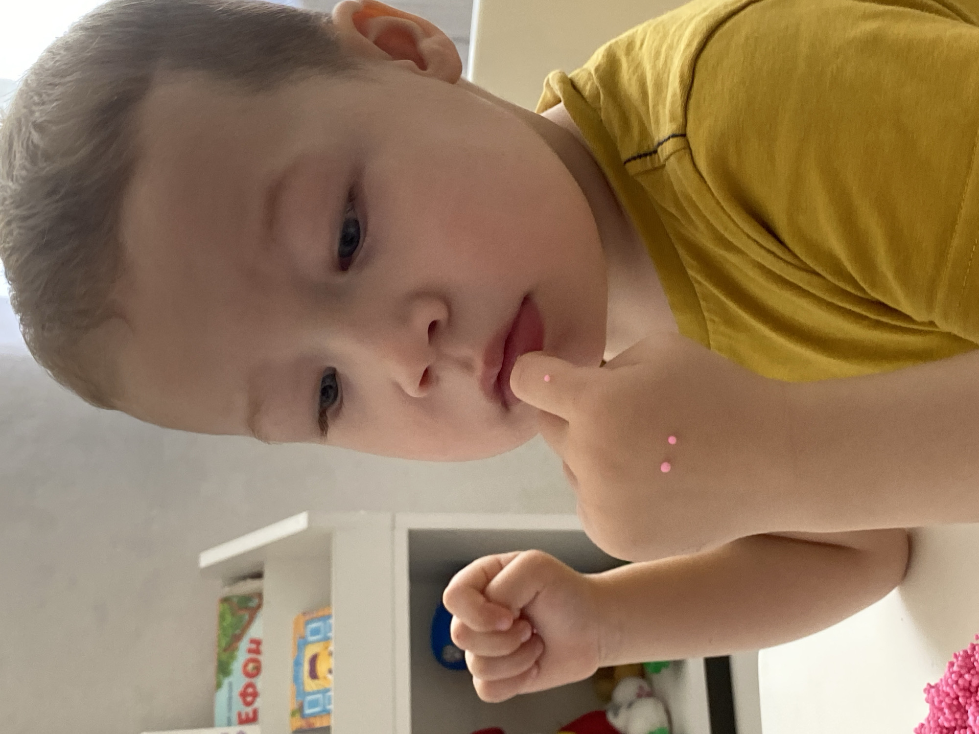 A young boy with fair skin and short, light brown hair is positioned at the top of the image, looking down at something. He is wearing a mustard-yellow t-shirt. His right hand is raised, with his finger near his mouth, and two small pink objects are adhered to his forearm. His left hand is clenched into a fist.
In the background, a white shelf is visible, containing books and toys. A table with pink material is also visible.
The information does not explicitly indicate the location is Sochi, Russia.