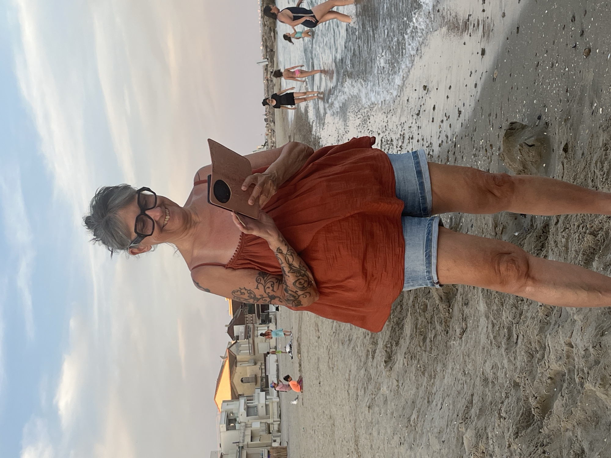 The image depicts a woman standing on a sandy beach in Palavas-les-Flots, France, holding a tablet. She is wearing a rust-colored tank top and denim shorts, with large-rimmed glasses. Tattoos are visible on her left arm. In the background, the Mediterranean Sea laps the shore, with several people, including children, wading or walking in the water and on the beach. Buildings typical of a coastal town are visible further inland. The sky is overcast with a pale, diffused light, suggesting it is either early morning or late evening. The scene captures a casual, everyday moment on a popular French beach.