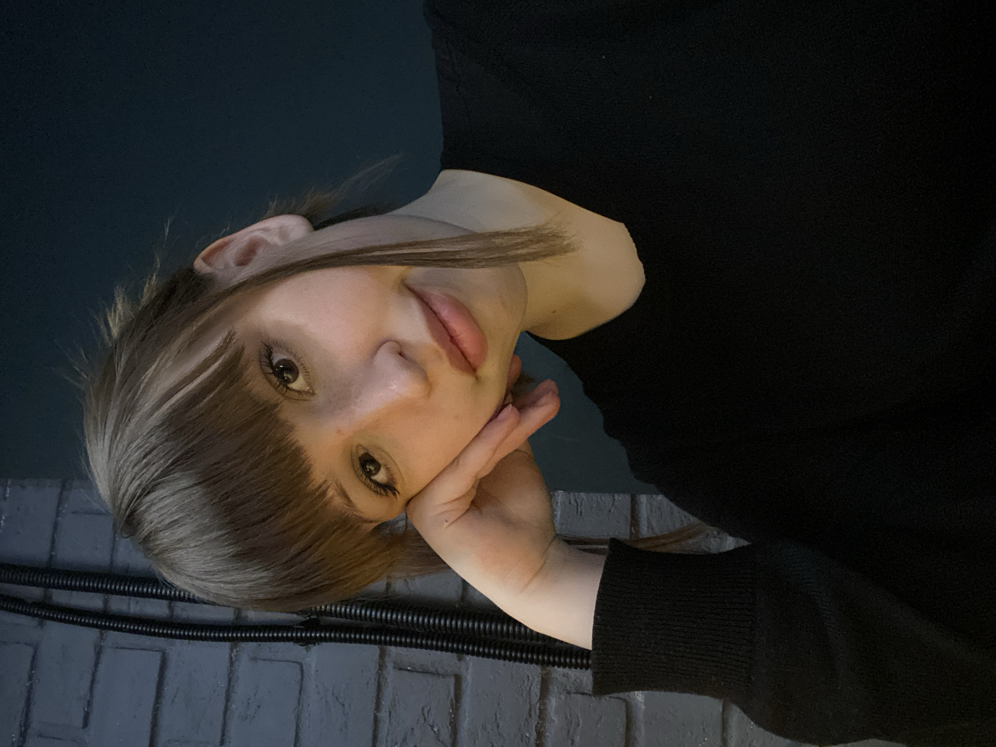 The image displays a close-up portrait of a young person, oriented sideways (rotated 90 degrees clockwise from a typical upright orientation). The subject is likely female, with light brown hair, bangs, and fair skin, featuring visible freckles on her nose. Her dark eyes are looking directly at the camera, and her expression is soft and contemplative. Her left hand is resting against her cheek and jawline, supporting her head in a relaxed pose. She is wearing a dark, possibly black, long-sleeved top or sweater.
The background is dimly lit, suggesting an indoor setting or a night-time scene in Chaikovsky, Russia. The upper portion of the background is a solid dark blue-green color, possibly a wall or a shadowed space. The lower half reveals a dark grey, textured surface, resembling brickwork or large tiles. Running horizontally across this textured surface are two prominent black corrugated pipes or cables, contributing to an industrial or functional aesthetic in the environment.
The overall atmosphere of the image is calm and reflective, emphasized by the subdued lighting and the subject's gentle gaze and posture. No visible text is present in the media file.