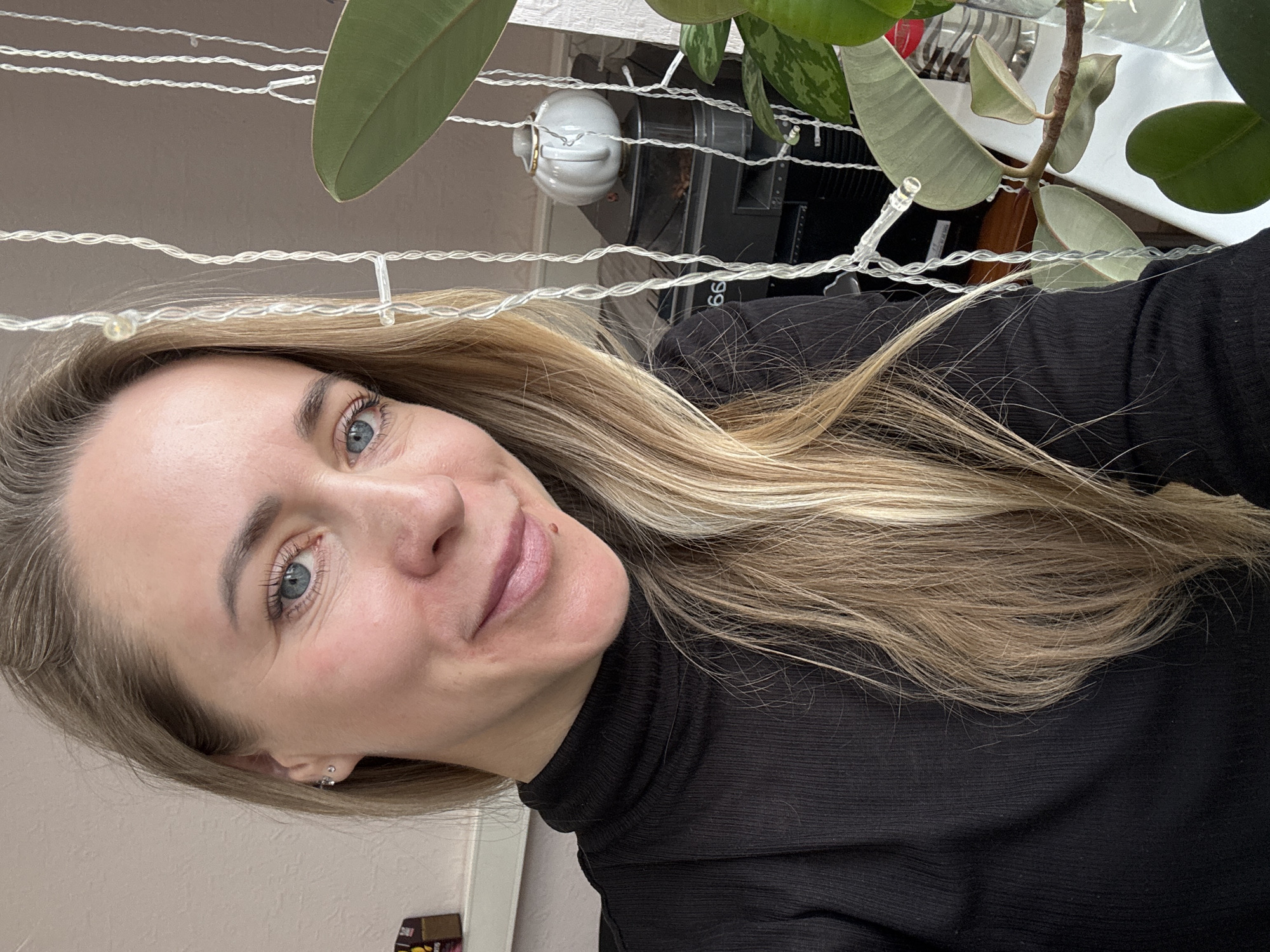 A close-up portrait features a woman with long, straight blonde hair, light blue eyes, and fair skin, captured indoors, likely in Chaikovsky, Russia. She displays a subtle, pleasant smile, with a small mole visible above her upper lip. She is dressed in a black ribbed turtleneck top. A silver stud earring is visible in her left ear.
The background features white string lights with small clear bulbs strung horizontally across the upper frame, adding a decorative touch. Large, dark green leaves of a houseplant, possibly a Ficus, are prominent in the upper and right sections, partially obscuring the deeper background.
Behind these foreground elements, several objects are partially visible. These include a small white ceramic teapot or sugar bowl with a gold handle, and dark electronic equipment, possibly a speaker or part of a computer setup, which bears the number "99". A faint red object and some white textured surfaces are also discernible. In the bottom left corner of the original image, a partial label or box with the red letters "SM" is visible. The woman appears to be posing for a close-up photograph or selfie, exuding a calm and friendly demeanor under indoor lighting.