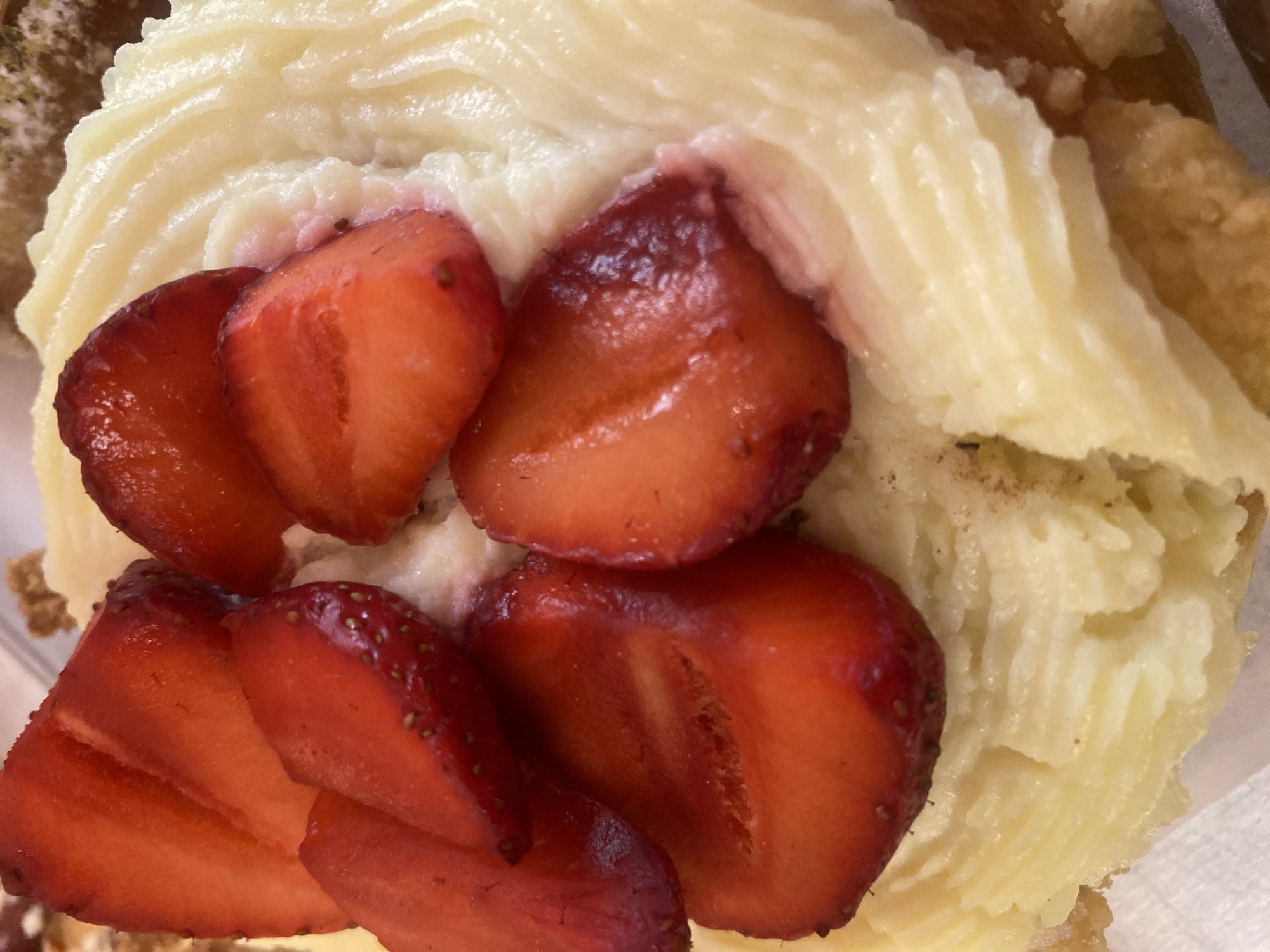 This is a close-up, overhead shot of a dessert, likely a cake or pastry, topped with cream and sliced strawberries. The cream is pale yellow and piped in a swirling pattern. Several slices of bright red strawberries are arranged on top of the cream, some showing their seeds. The texture of the cream appears smooth and rich, while the strawberries are juicy and fresh. The dessert sits on a white surface, possibly a plate or napkin. The lighting suggests it could be indoors, with natural light contributing to the bright, appealing appearance of the food. There are no people or discernible textual elements in the image. The overall impression is one of a delicious and inviting sweet treat.
