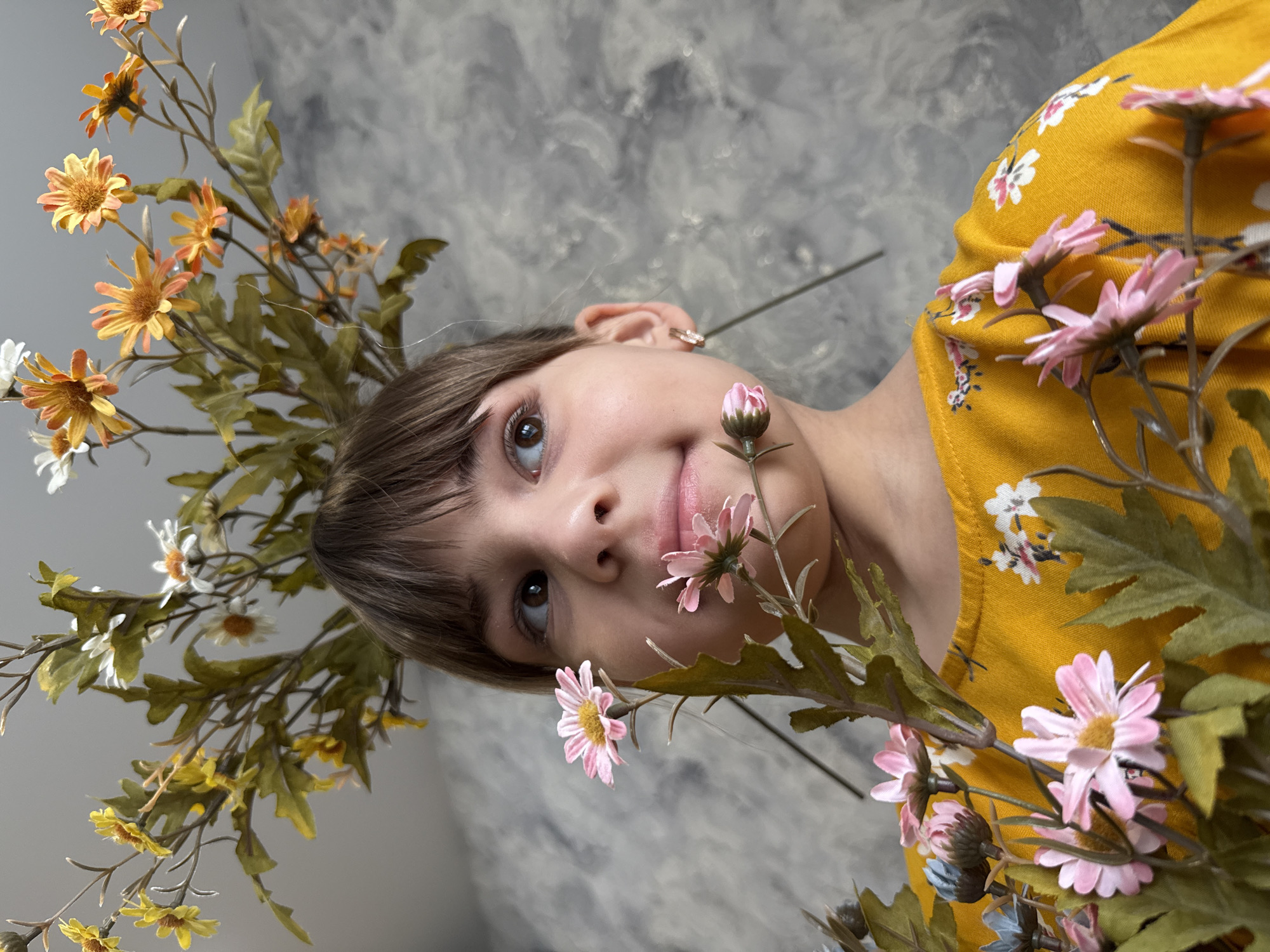 A close-up, tilted shot features a young person with light skin and brown hair, adorned with artificial flowers in shades of yellow, orange, pink, and white with green foliage. The person is wearing a mustard-yellow top with a floral pattern. The background is a mottled grey, possibly a wall. The setting appears to be indoors. The location is Unknown, Unknown.