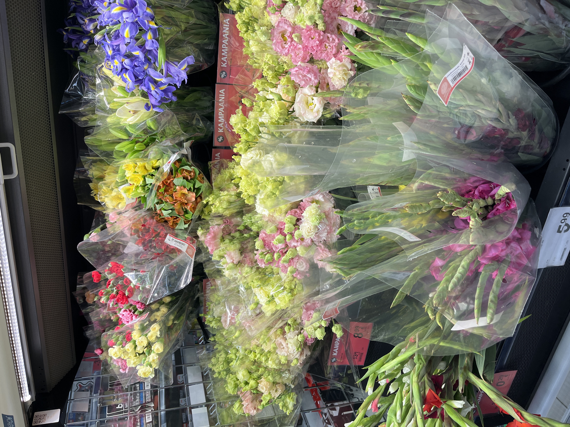 This is a medium-shot, eye-level, full-color, vertically oriented photograph of a flower display at a retail establishment in Kohtla-Järve linn, Estonia. The flowers are arranged in bouquets and wrapped in clear plastic. The display is sectioned into multiple shelves. On the top shelf, there are bouquets of various colors including pink, yellow, red, and orange flowers. The second shelf from the top features a bouquet of purple iris flowers. The third shelf is filled with bouquets of pink and light green flowers. The bottom shelf displays bouquets of gladiolus flowers in pink and red hues. Red and white price tags are visible on some of the bouquets, with text that appears to be in Finnish or Estonian. The lighting appears to be from overhead fluorescent lights. There are no people visible in the image.
