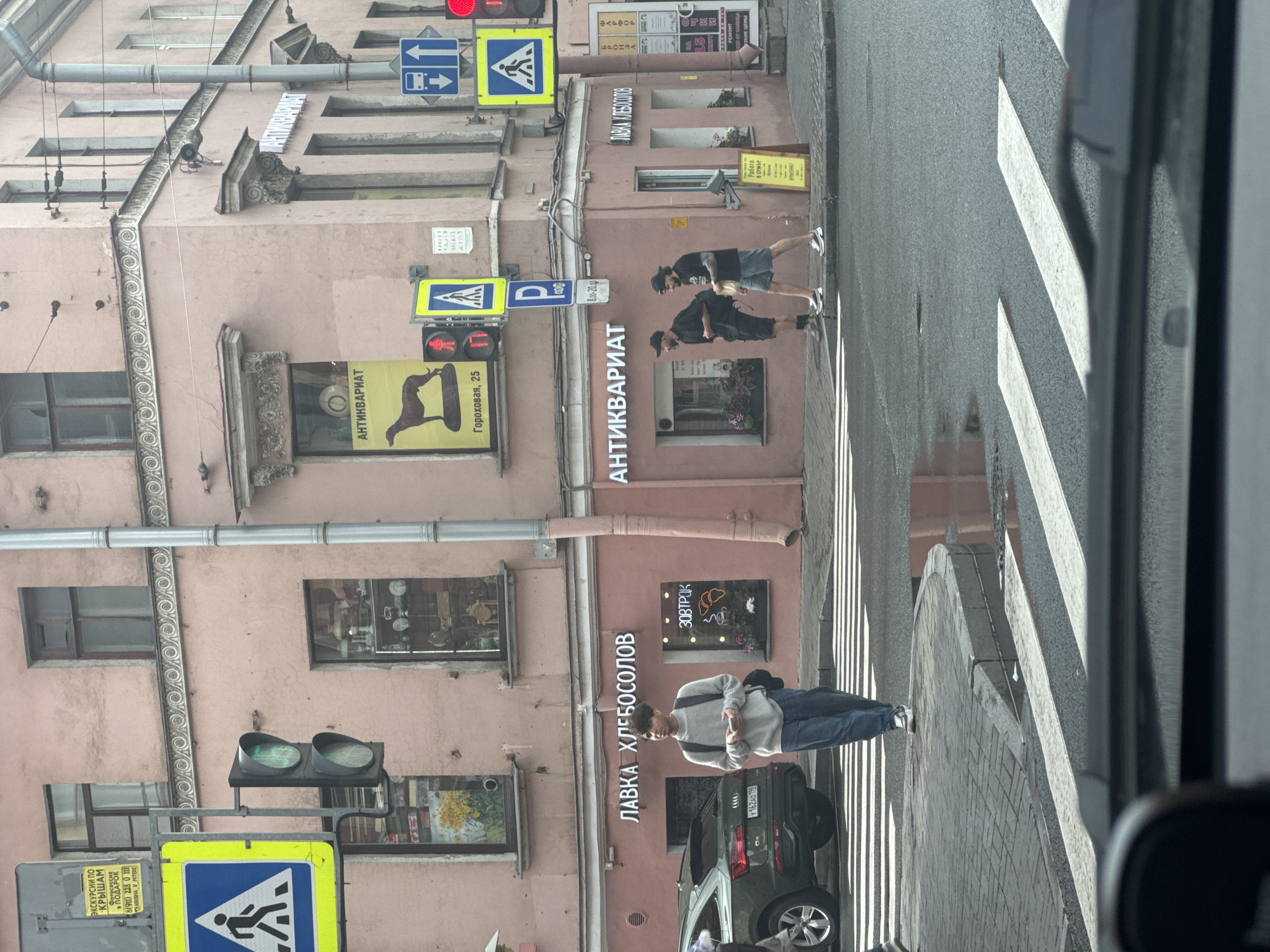 The image captures a street scene in Saint Petersburg, Russia, focusing on a building with shops and pedestrians crossing a street. The building, painted a pale pink, features architectural details like ornate moldings around windows and along the facade. Two storefronts are visible: one labeled "ЛАВКА ХЛЕБОСОЛОВ" (Lavka Khlebosolov, meaning "Bread-Lovers' Shop") and another with the sign "АНТИКВАРИАТ" (Antikvariat, meaning "Antiques"). A yellow sign with a silhouetted dog and text "Гороховая, 25" (Gorokhovaya, 25) is displayed near the antique shop.
Several traffic lights and street signs are present. A pedestrian crossing is marked on the asphalt, and there are puddles of water on the street, suggesting recent rain.
In the foreground, a young man with dark hair, wearing a light grey sweater and blue jeans, is walking across the pedestrian crossing, holding a smartphone. To his right, two men are also crossing the street: one in a black t-shirt and shorts, and the other in a dark blue t-shirt and shorts. A dark green SUV is parked to the left of the pedestrian crossing.
The overall atmosphere suggests a typical urban street environment in Saint Petersburg.