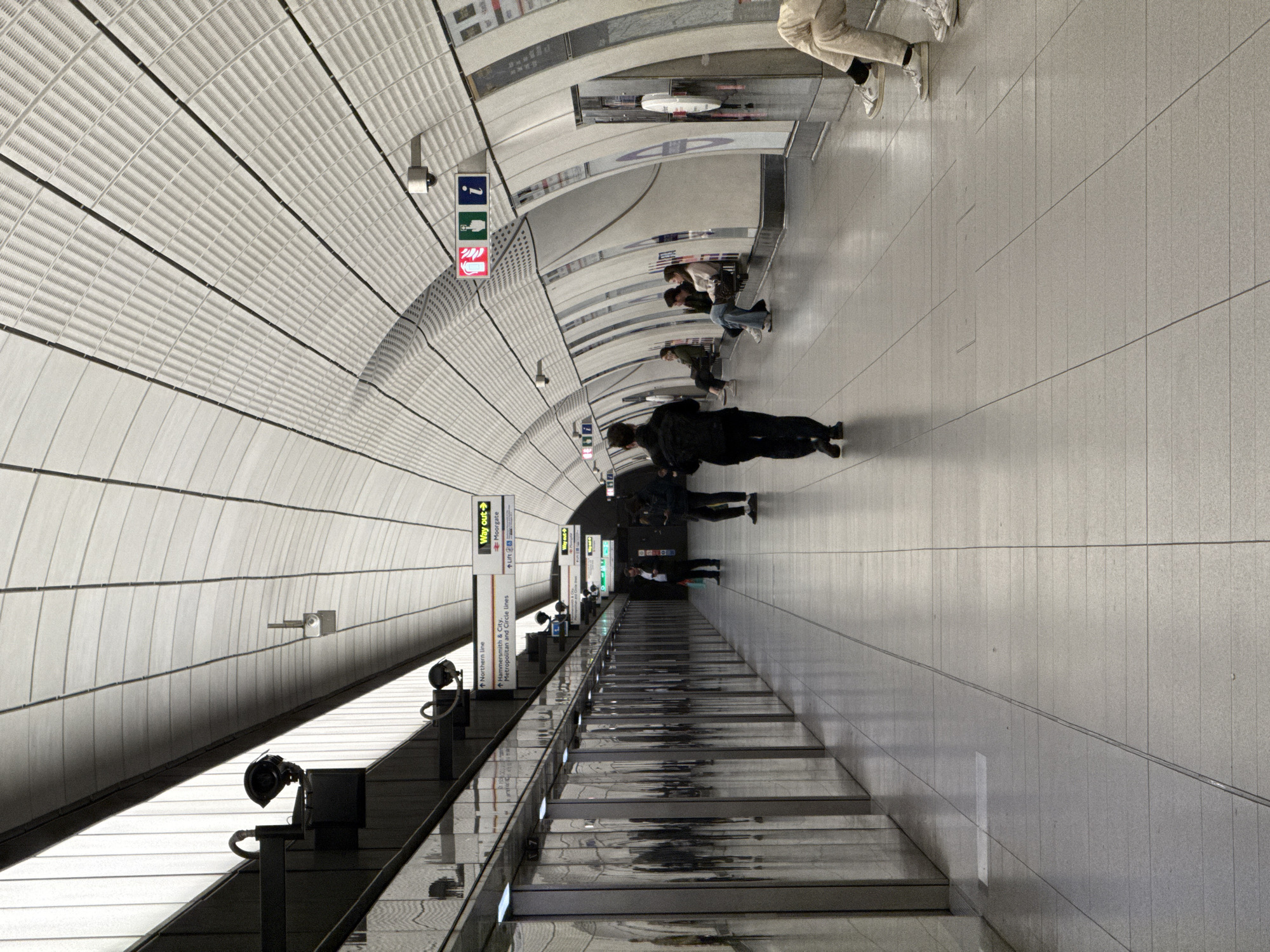 Certainly, here's a detailed description of the image:
**Scene:** The image depicts a section of a London Underground (Tube) station. It's shot at an angle, emphasizing the perspective of the platform stretching into the distance. The station appears modern and well-lit with a white color scheme dominating the scene.
**Environment:** The ceiling is curved and paneled, likely for acoustic or aesthetic purposes. Walls are tiled in light grey. There are stairs leading up.
**People:** Several people are visible on the platform. Some are walking, while others are seated on benches. There's a group of people towards the middle ground of the image.
**Objects:** There are directional signs indicating "Way Out," "Moorgate," and "Lift". Other signs note the "Northern Line" and the "Hammersmith & City, Metropolitan and Circle Lines." Lighting fixtures are mounted on the walls and ceiling. Railings line the edge of the stairs.
**Inferences:** The overall impression is that of a modern, functional transportation hub that serves a busy urban population. Based on the signage, the location is Moorgate Station on the London Underground.