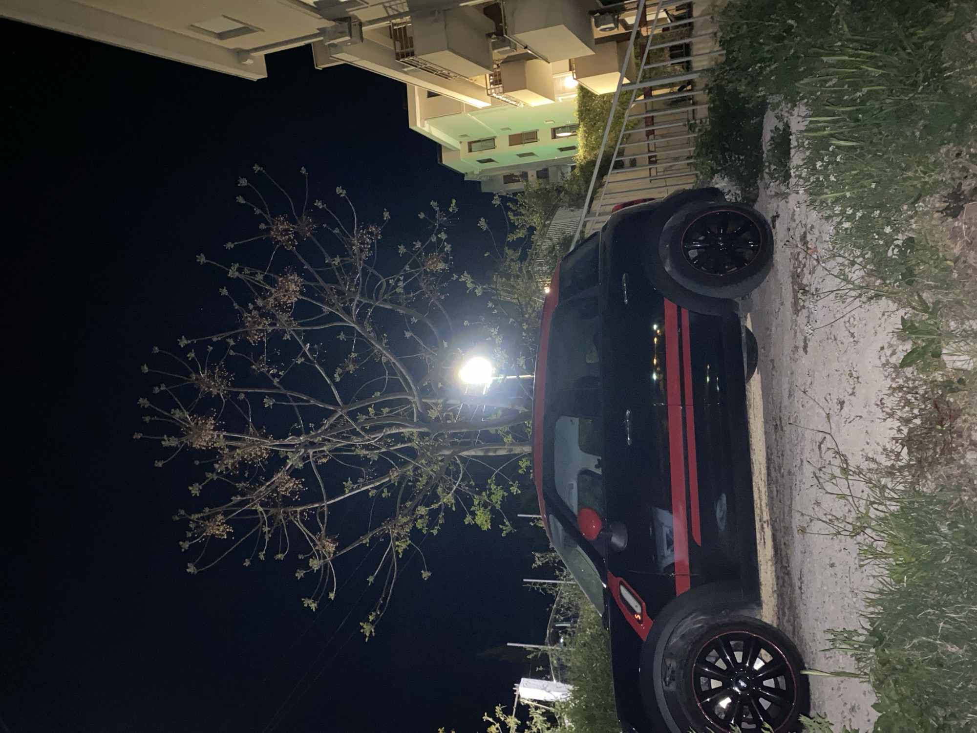 A black car with red racing stripes is parked on a narrow sidewalk near a building at night. The building is light colored and multi-story. A tree is adjacent to the car, positioned under a street lamp, which illuminates the surrounding area. Vegetation is growing on the right side of the frame. The location is Budva, Montenegro.