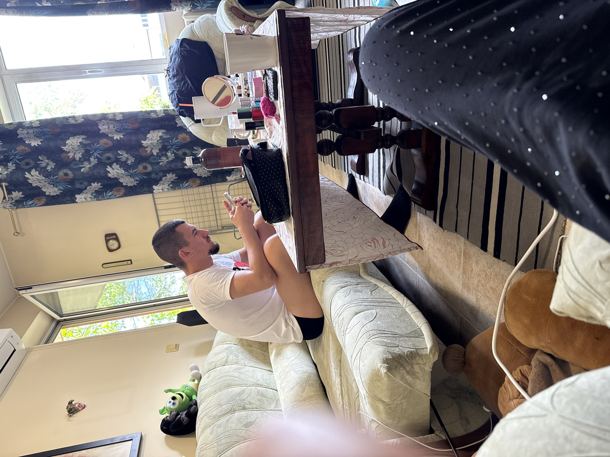 A person is sitting cross-legged on a patterned, light-colored sofa, using a mobile phone. The person is wearing a white t-shirt and black shorts. A wooden table is partially visible in the frame, with makeup items, a mirror, and a black purse on it. The room features floral-patterned curtains and a window showing trees outside. A black and white striped rug is visible on the floor. On the wall there is a clock, a air conditioner, a framed photo and a soft toy. The image may be taken in Sofia, Bulgaria, if that is the location. The scene is indoors, showing a snapshot of casual domestic activity.