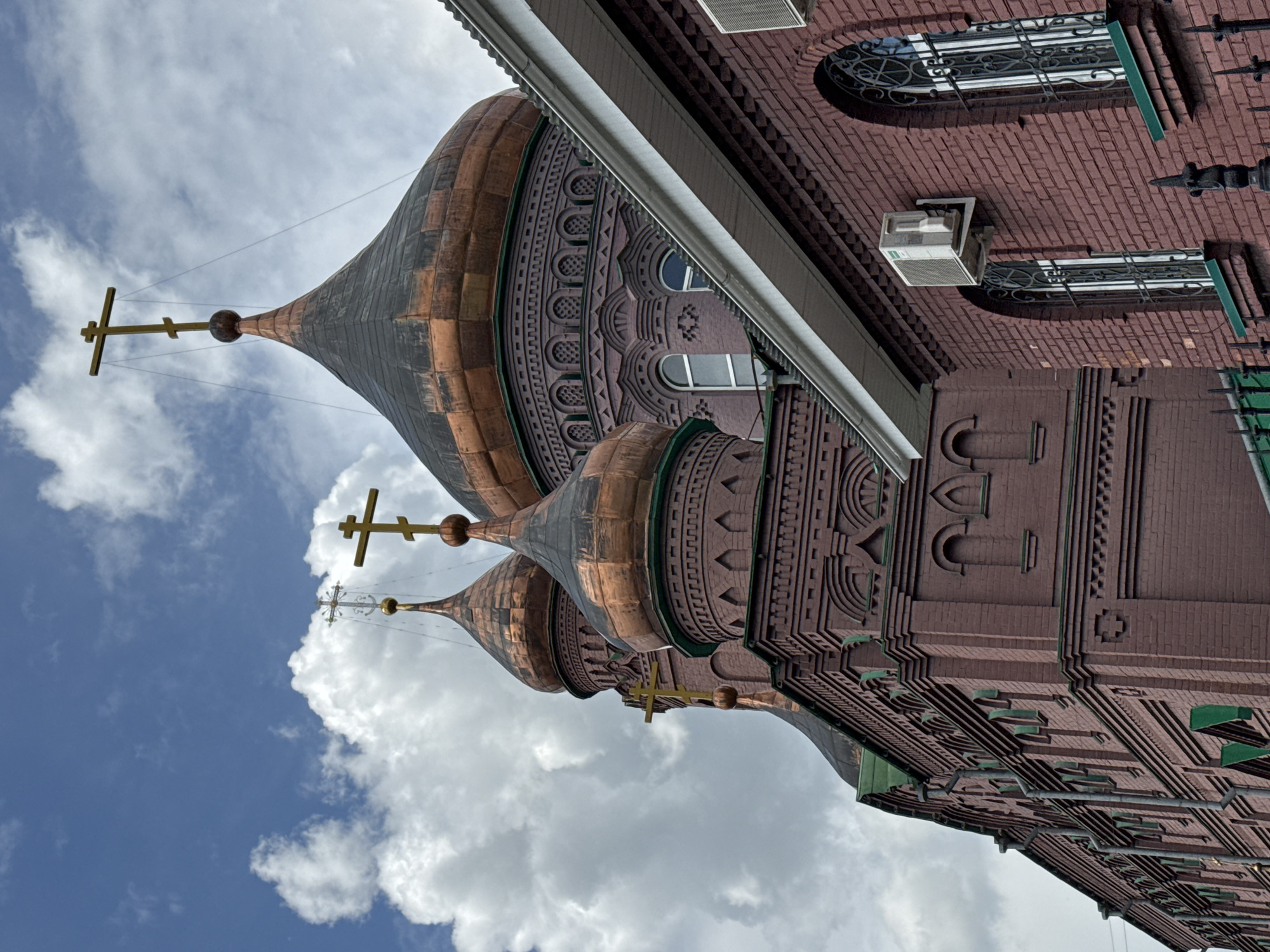 An upward-angled shot captures the ornate facade and distinctive domes of a red-brick building, likely a church or a historically significant structure, set against a partially cloudy blue sky. Three onion-shaped domes, seemingly constructed from patinated copper or bronze, rise prominently on the left side, exhibiting a mottled texture of brown, black, and green hues. Each dome is crowned with a golden Orthodox cross.
The building's facade is made of dark red brick, intricately adorned with decorative elements and patterns carved or molded directly into the brickwork. These details are visible in the arched recesses, geometric designs, and stylized motifs running along the upper sections and beneath the domes. Several windows punctuate the wall; some are arched with ornate iron grilles, while others are rectangular with simple white frames. A white, rectangular outdoor air conditioning unit is mounted on the brick wall, positioned between two windows.
The sky behind the structure features patches of clear blue interspersed with soft, white cumulus clouds, indicating a bright daytime setting. The perspective emphasizes the height and architectural detail of the edifice. No visible text is present in the image.